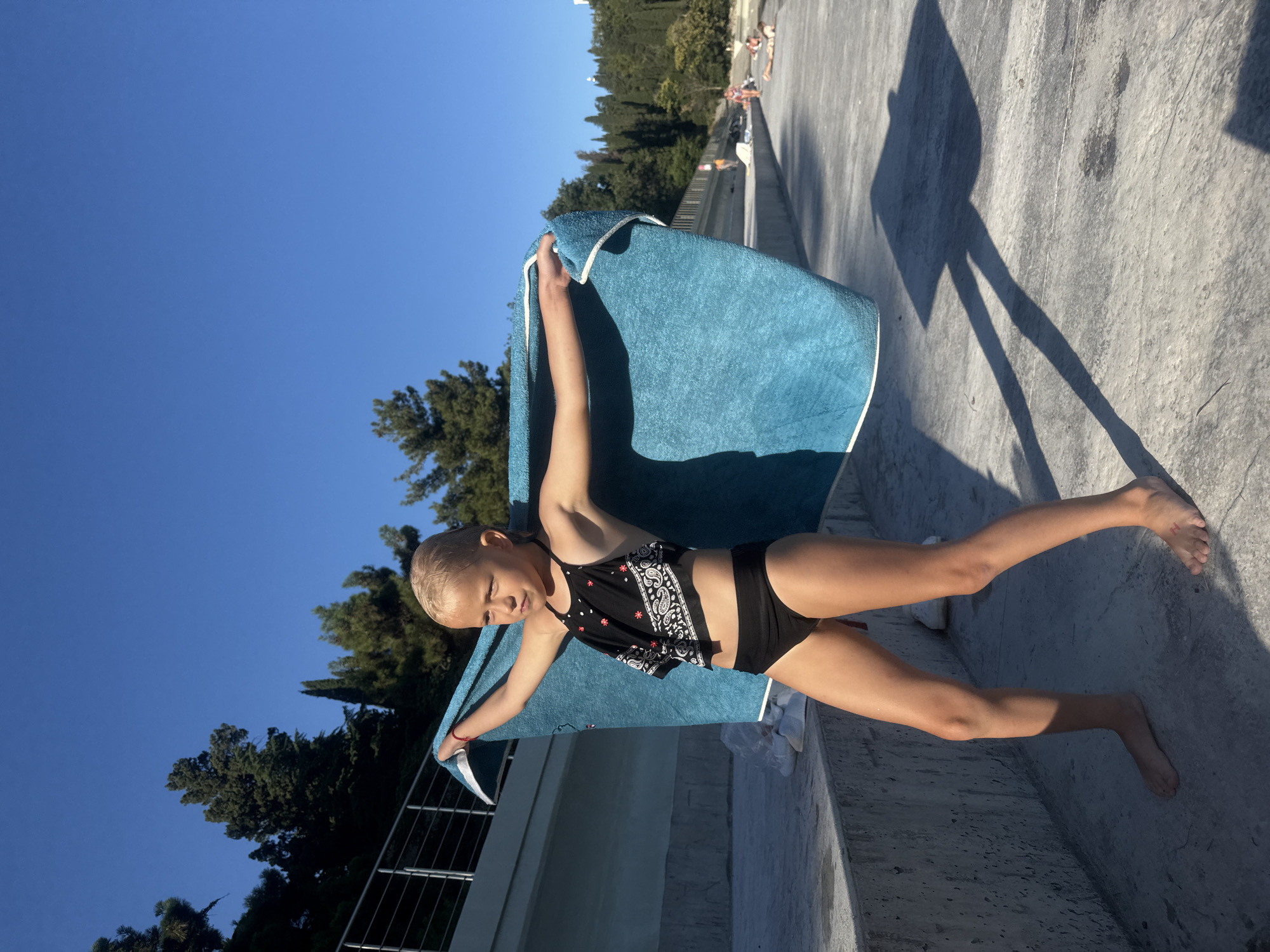 A young girl in a black swimsuit stands outdoors, holding a bright blue towel. She is in a dynamic pose, with her arms outstretched and one leg extended. Her facial expression is one of playful confidence. The setting appears to be a concrete area with a railing, possibly near a pool or a viewing platform, given the presence of other people in the background relaxing on lounge chairs.
The time of day is evident from the long shadows cast on the concrete, suggesting it is late afternoon or early evening. The sky is a clear, vibrant blue, indicating fair weather. Lush green trees are visible in the background, adding a natural element to the scene.
The girl's blonde hair is short and swept back. The swimsuit has a black and white patterned top and plain black bottoms. The blue towel she holds has white edging. On her right foot, a small red mark that looks like an "H" is visible. There is no visible text in the image. The overall impression is one of a sunny day outing, with the girl enjoying herself.