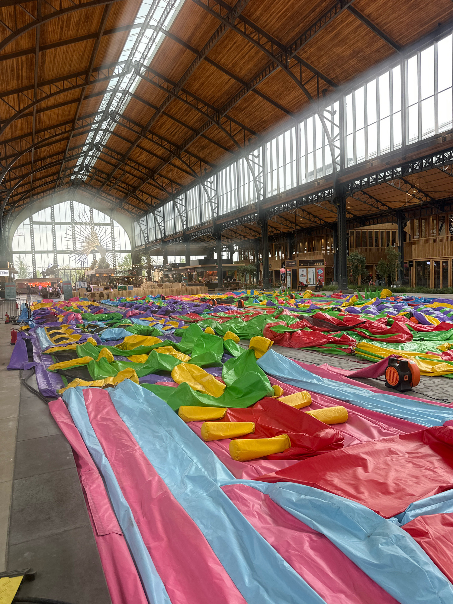 The image depicts a large, indoor space with a high, wooden-beamed ceiling and extensive glass windows. The floor is covered with a vast expanse of deflated, brightly colored inflatable structures, predominantly in shades of pink, blue, yellow, green, and red. These inflatables are folded and spread out, suggesting they are either being set up or taken down. In the background, the space appears to be a public area or venue, with some people visible, seating, and food stalls or shops. An orange blower fan is situated on the inflatable material in the foreground, likely used to inflate the structures. The scene is set in Brussels, Belgium, at the historic Gare Maritime, a former train station now transformed into a commercial and event space.