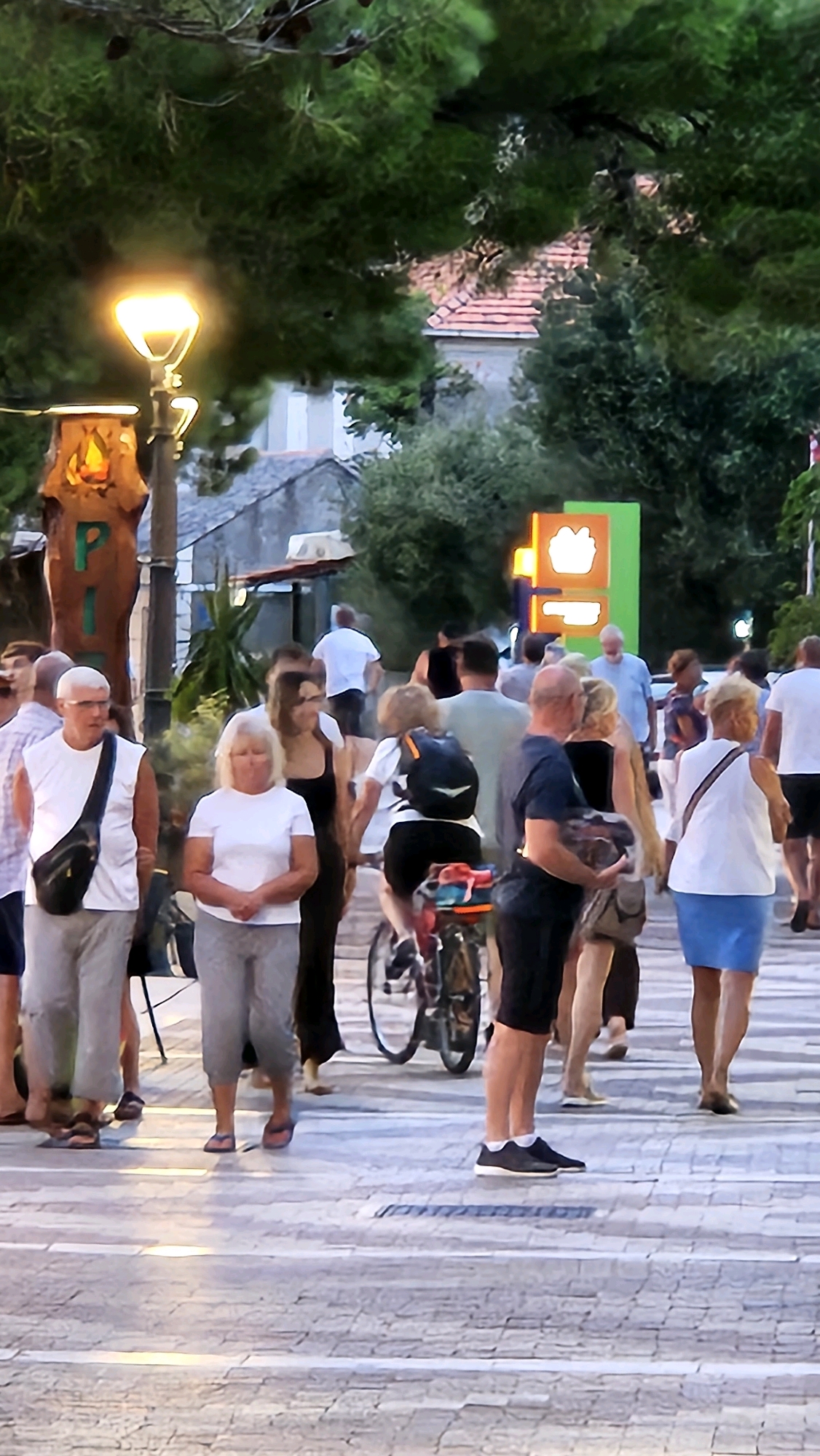 In Općina Tučepi, Croatia, a street scene unfolds as a group of people walk across a paved pedestrian crossing. The time of day appears to be late afternoon or early evening, suggested by the warm glow of a street lamp and the soft lighting filtering through the trees.
In the foreground, several individuals are captured in mid-stride. A man in a white t-shirt and grey pants, with a black cross-body bag, is walking from left to right. Next to him, a woman in a white t-shirt and grey cropped pants is also walking, looking downwards. Behind them, a person in a black tank top walks, followed by another woman in a white t-shirt and a blue skirt, carrying a bag over her shoulder.
Further into the scene, a cyclist on a red bicycle is navigating the crossing. Other pedestrians are visible in the background, some walking and some standing. The environment is characterized by a paved street with white lines marking the pedestrian crossing. Lush green trees overhang the scene, with hints of buildings and an illuminated sign with orange and yellow elements visible in the distance. The overall impression is that of a lively, public space with people enjoying a casual stroll.