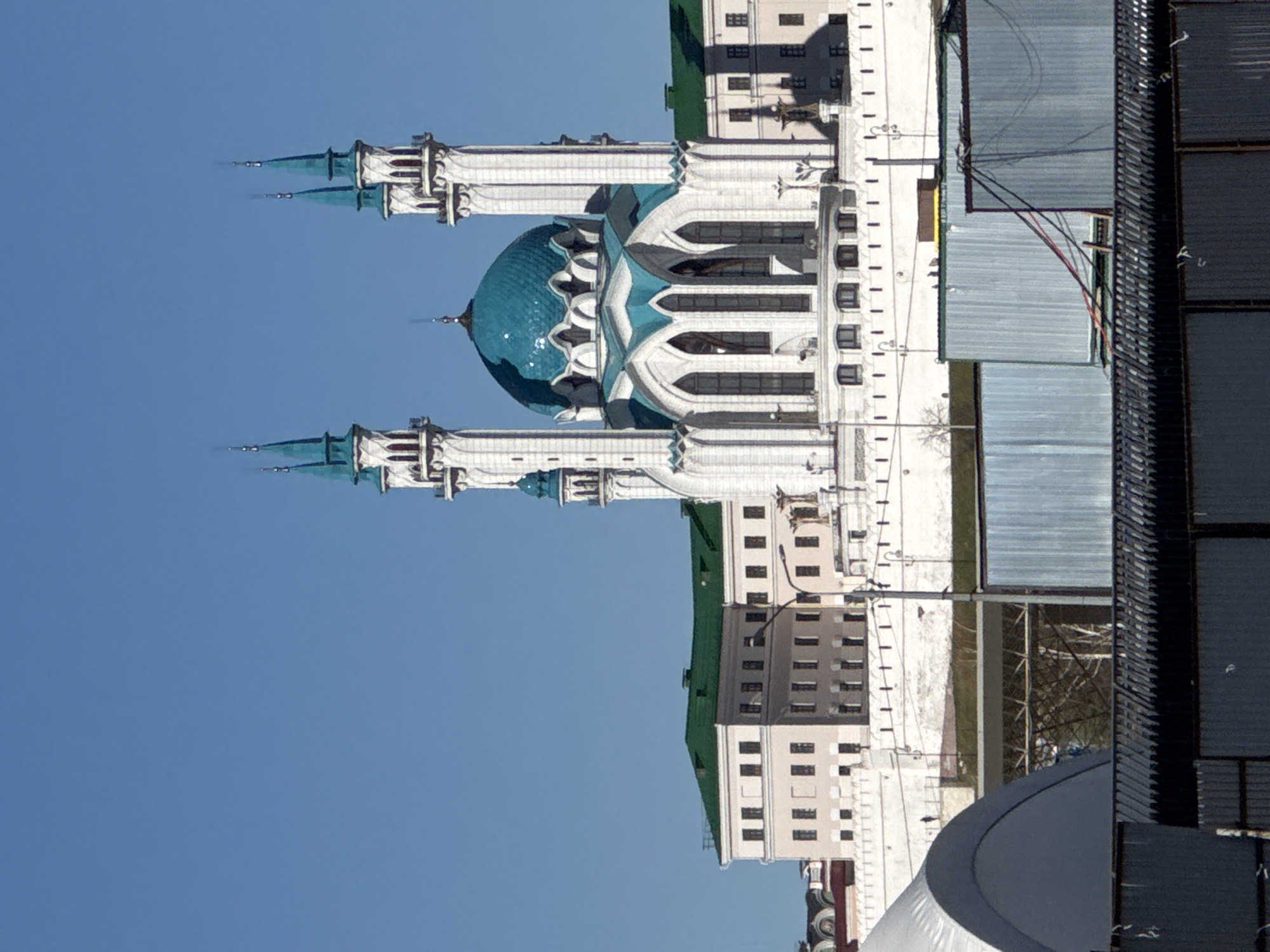 The image shows the Qolsharif Mosque in the Kazan Kremlin, Kazan, Russia. The mosque, with its blue dome and minarets, dominates the scene against a clear blue sky. A building is adjacent to the mosque. Other buildings are visible at the edge of the image.