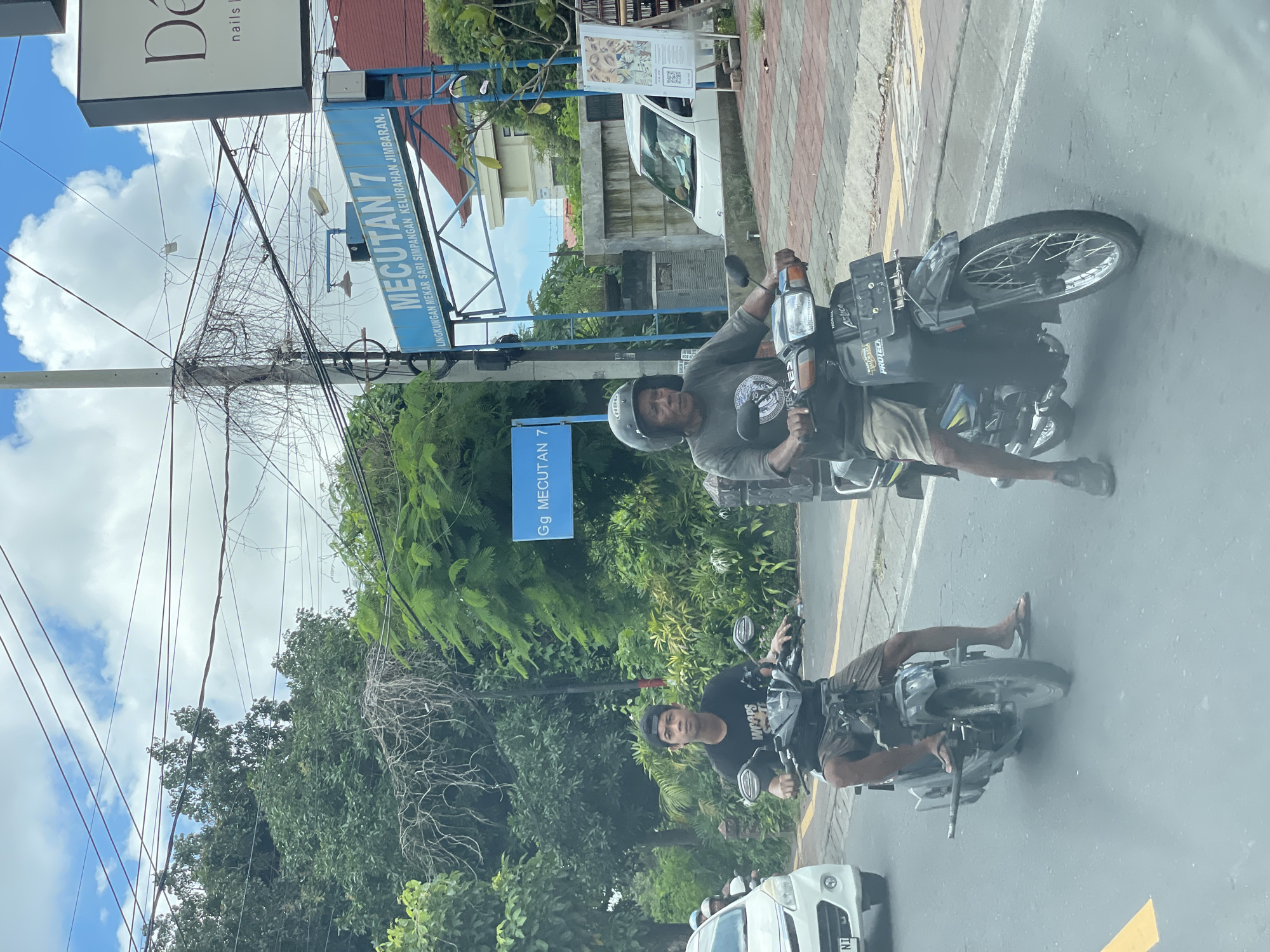 Two men ride motorcycles on a street in Kuta Selatan, Indonesia. The man on the front motorcycle wears a helmet, grey shirt, and khaki shorts. The other man wears a black t-shirt and hat. Signage indicates the location is Gg Mecutan 7 and Mecutan 7, with a longer inscription reading: LINGKUNGAN MEKAR SARI SIMPANGAN KELURAHAN JIMBARAN. Overhead, a tangle of electrical wires stretches across the sky near a "De Nails" sign. A white car follows behind the second motorcycle. The scene is partly obscured as if viewed through a car windshield.