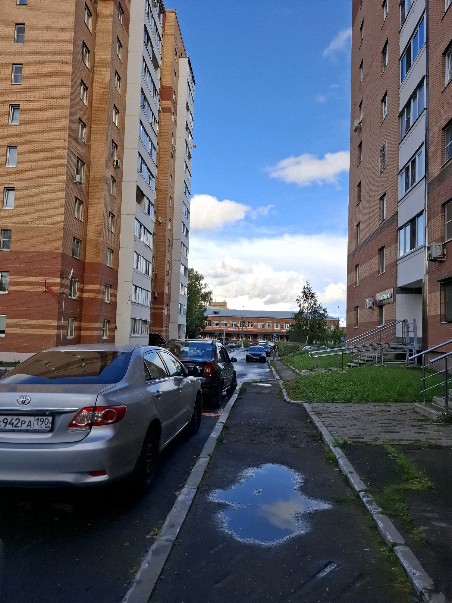 This is a medium-shot, eye-level, vertical, outdoor photograph taken in a city environment. The scene is dominated by two tall, multi-story apartment buildings constructed with red brick and white trim, positioned on opposite sides of a paved walkway and road. The sky is blue with scattered white clouds.
In the foreground, a silver Toyota Corolla sedan is parked on the left side of the frame. Its license plate reads "C942PA 190". To the right of the Corolla, a black sedan is also parked. The walkway in the center of the image is wet, with a large puddle reflecting the sky and clouds. Beyond the walkway, a strip of green grass separates it from the sidewalk in front of the building on the right.
The building on the left features numerous windows and air conditioning units mounted on its facade. A small red flag is visible on this building. The building on the right also has many windows and balconies. Stairs with metal railings lead up to an entrance on this building, where a sign with Cyrillic lettering is visible. Further in the distance, more apartment buildings and trees can be seen. The overall impression is a residential area, possibly after a recent rain.
