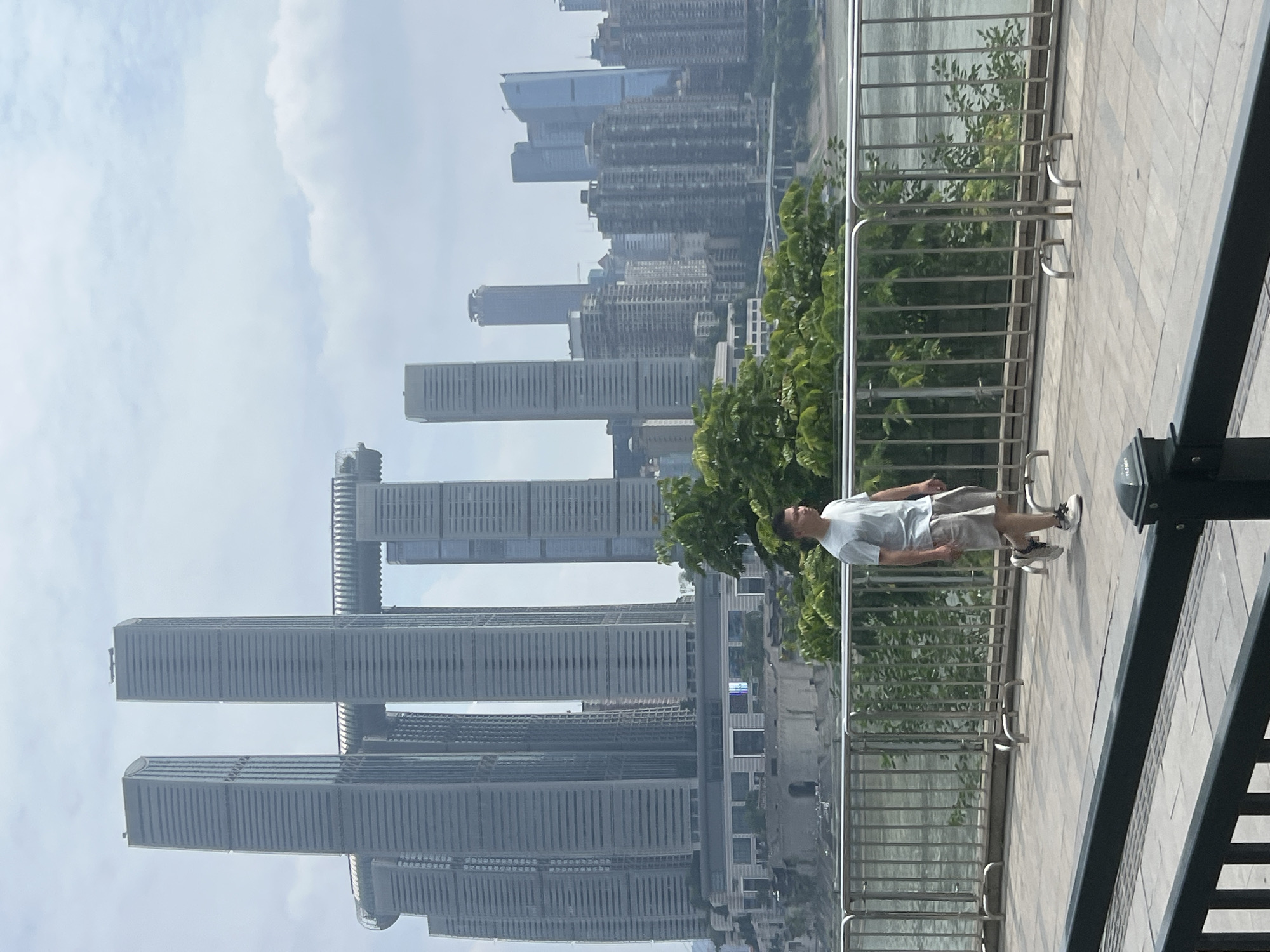 A man walks on a paved walkway in front of a metal railing with a river and a dense urban skyline in the background. The man, who appears to be East Asian, is wearing a light blue t-shirt, light-colored shorts, and white sneakers. He is walking to the right, looking straight ahead. Behind him, the walkway is bordered by a metallic railing and lush green trees. The river beyond the railing is wide and reflects the overcast sky. The skyline is dominated by modern skyscrapers of various heights and designs, with one notable building complex featuring multiple tall towers connected by a horizontal structure. The architectural style suggests a major city, likely in Asia, given the context provided about Nan'an District, China. The sky is cloudy, and the overall lighting suggests daytime. The foreground shows another, lower railing and a portion of the paved walkway.