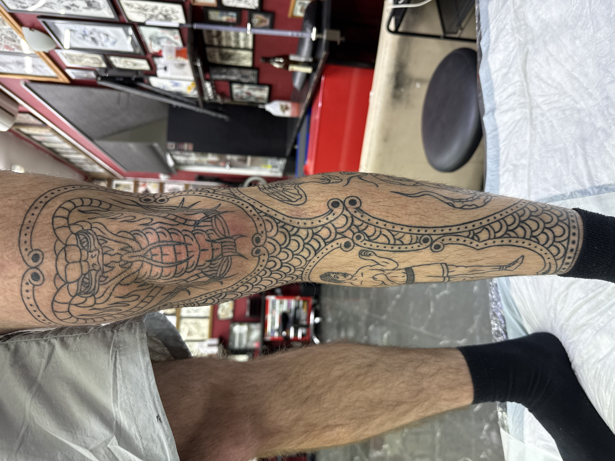 A person's lower leg and thigh are prominently featured, adorned with intricate, black-line tattoos. The leg is positioned horizontally, suggesting it's resting on a surface, with the foot partially covered by a black sock. Light-colored shorts are visible on the thigh.
The tattoos cover a significant portion of the leg, showcasing a detailed design that includes a large, scaled serpent or dragon-like creature around the knee area, with its head and part of its body visible. Further down the calf, the design incorporates repetitive scale patterns, decorative borders with circular and dotted motifs, and a human-like figure with a fish tail, possibly a merman or mermaid. The tattoos are rendered exclusively in black outline, suggesting they are either freshly completed linework or awaiting further shading and coloring.
The scene appears to be a tattoo studio or parlor. In the background, a wall is visible, densely covered with numerous framed pictures or artwork, likely tattoo flash. A red cabinet or shelving unit and a black, circular stool are also partially visible, along with a white, disposable medical drape on a table or bed surface. The general lighting suggests an indoor setting. This image is set in Moscow, Russia. No discernible text is visible within the image.
