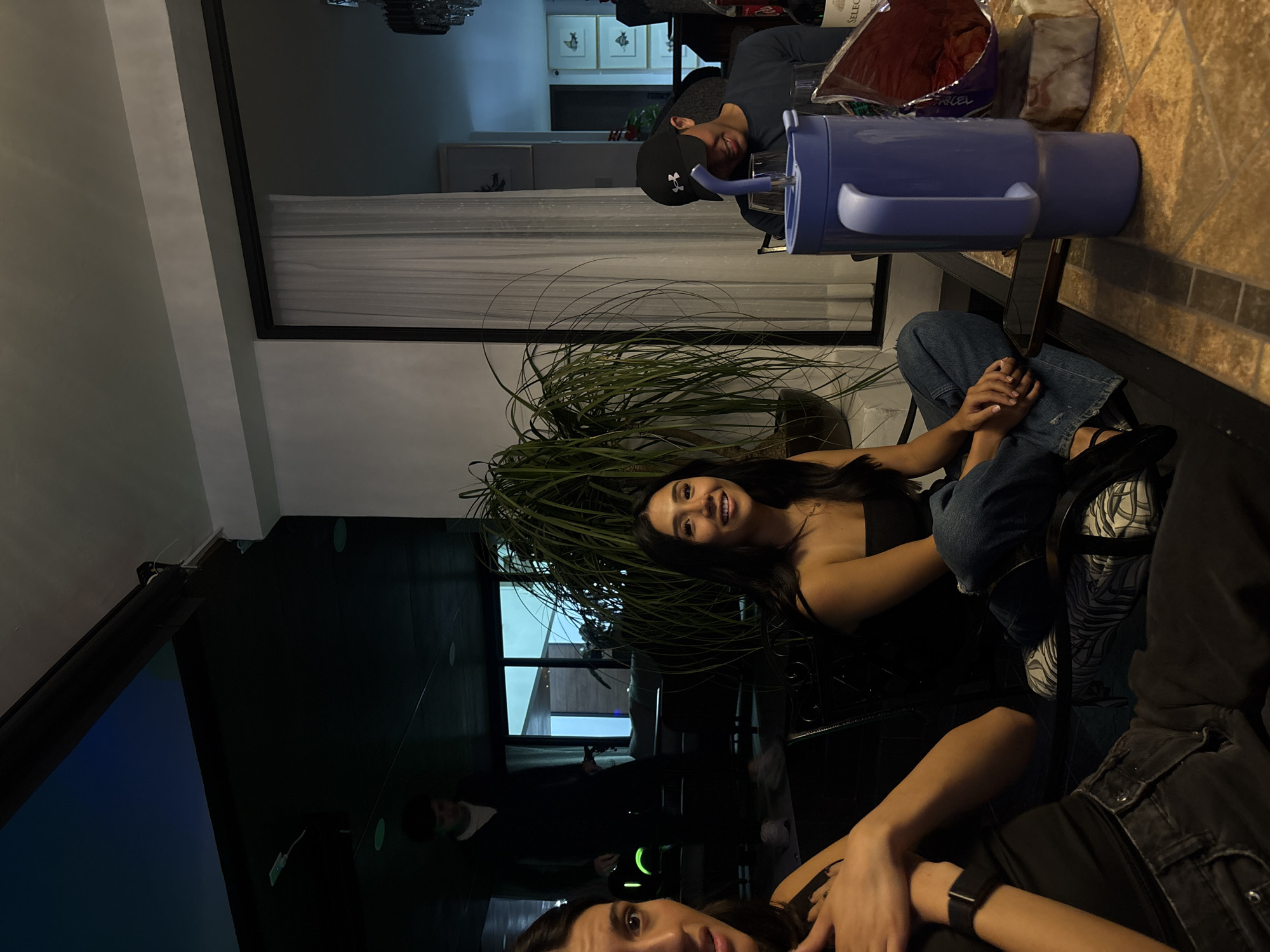 The scene depicts an indoor social gathering within a residence in San Bernardino Tlaxcalancingo, Mexico. Three individuals are prominently featured, with a fourth person in the background.
On the left, partially cropped, a woman with dark hair wears a dark top. Her left arm is raised, and a dark smartwatch is visible on her wrist. Her expression is one of mild surprise or concern, with her mouth slightly open and eyes wide, directed leftward out of frame.
In the center, a woman with long dark hair is seated, smiling and looking slightly to the right of the camera. She wears a dark strapless top and blue jeans, sitting cross-legged on a dark chair. Her hands are clasped in her lap.
To the right, partially visible behind a large purple insulated mug, a man in a dark baseball cap (featuring an Under Armour logo) and a dark shirt is seated. He is looking downwards or away from the camera, with a faint smile.
In the background, on the left side of the image, another man is walking through a dimly lit area, wearing a dark sweater and light-colored shoes. A glowing green object, possibly a speaker or illuminated toy, rests on the dark floor near his feet.
The environment suggests a living or dining area. A large potted plant with numerous long, slender green leaves stands between the two women and the man on the right. The furniture includes dark chairs and a table with a tiled surface in the foreground on the right. On this table are clear glasses, a partially consumed bag of chips or snacks, a smartphone, and a bottle of red wine labeled "SELECCION".
The lighting is subdued, with some light sources from overhead (recessed lights are visible in the ceiling) and from a large window or glass door on the right, which is covered by sheer white curtains. Beyond this window, framed artwork, possibly depicting fish, hangs on a distant wall. Overhead on the far right, a crystal chandelier is partially visible. The left side of the room features darker walls and ceiling elements, possibly indicating a different section or a covered outdoor patio area.