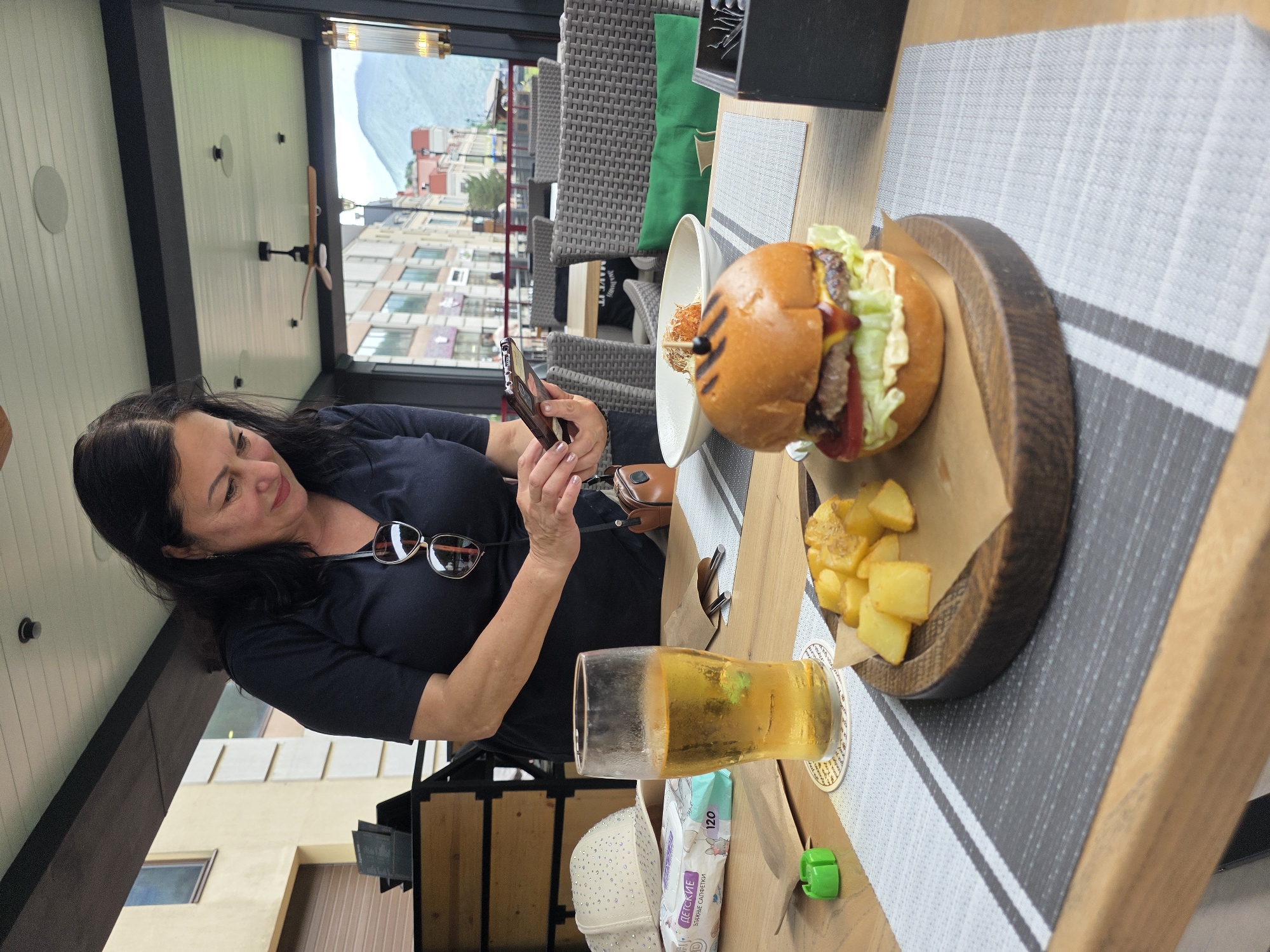In Estosadok, Russia, a woman with dark hair, wearing a dark blue short-sleeved shirt, stands by a table set with food and drinks. She is looking down at a smartphone she holds in her hands. On the table in front of her are a glass of light-colored beer, a burger with fries, and a package of wet wipes. The scene appears to be an outdoor dining area with a covered roof and wicker chairs visible in the background. The woman is wearing sunglasses on top of her head.