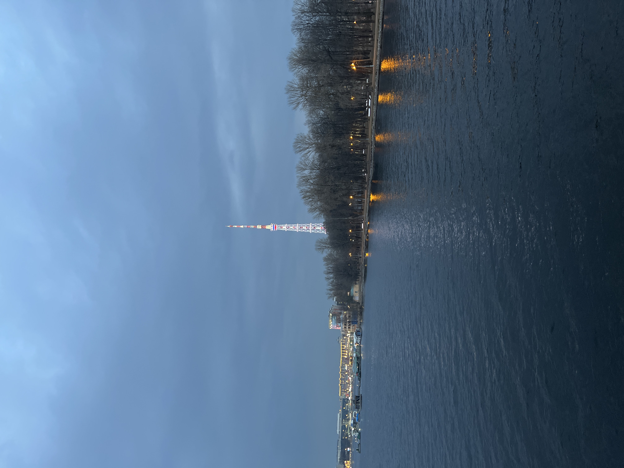 The image depicts an urban waterfront scene in Saint Petersburg, Russia, observed during twilight or early evening under an overcast, dark blue-grey sky. The foreground is occupied by the dark blue, rippled surface of the Neva River, which reflects the amber glow of streetlights.
Along the right bank, a tree-lined embankment, identified as Petrovsky Park, extends horizontally. It is densely populated with bare deciduous trees, indicating a dormant season. Multiple evenly spaced streetlights cast orange illumination onto the embankment and reflect in the river. Several white park benches are discernible along the path adjacent to the water.
In the mid-ground, centered above the tree line, the Saint Petersburg TV Tower stands prominently, its lattice structure illuminated with vertical bands of red, white, and blue lights, resembling the Russian flag.
Further to the left, across the river, a cluster of brightly lit buildings and structures forms part of the city's infrastructure. This includes a small marina or dock area with several boats or floating platforms. No human figures, vehicles, or specific activities are discernible within the frame.