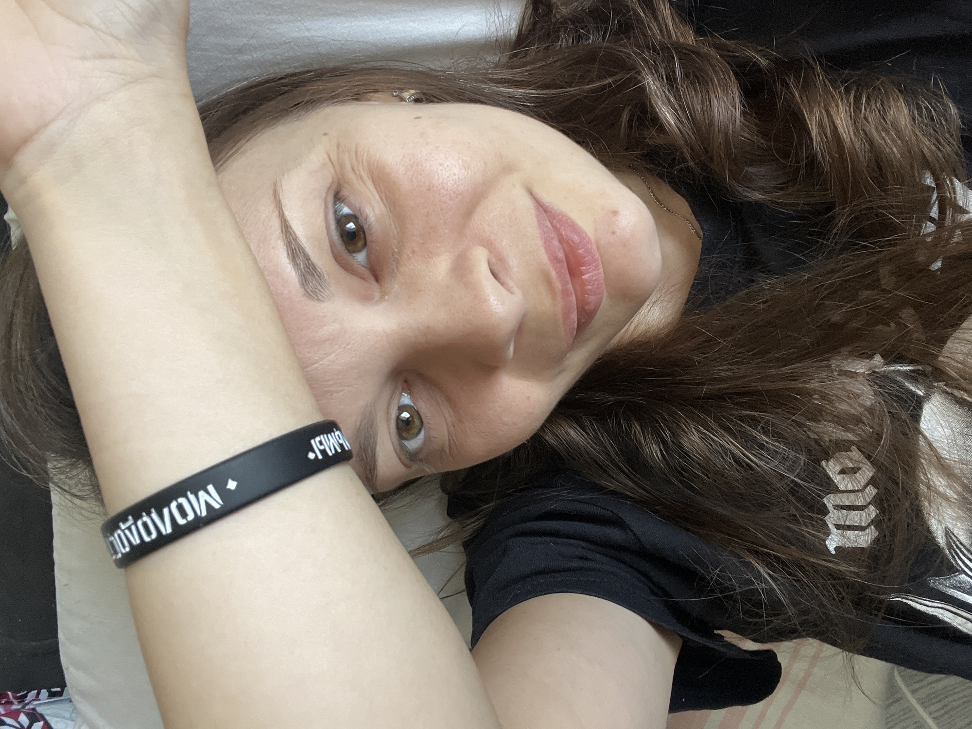 The image shows a close-up of a woman's face and arm, with her hand resting on her forehead. She is wearing a black t-shirt and a black wristband with white lettering that appears to say "Молодежь" (Molodezh), which means "youth" in Russian. The woman has brown hair and brown eyes, and she is smiling slightly. Her skin shows signs of natural aging, with fine lines around her eyes. A delicate necklace is visible around her neck. The background is a plain white surface, possibly a bedsheet, suggesting an indoor setting. There is no indication of the city of Tolyatti, Russia, within the image itself or in any provided context.