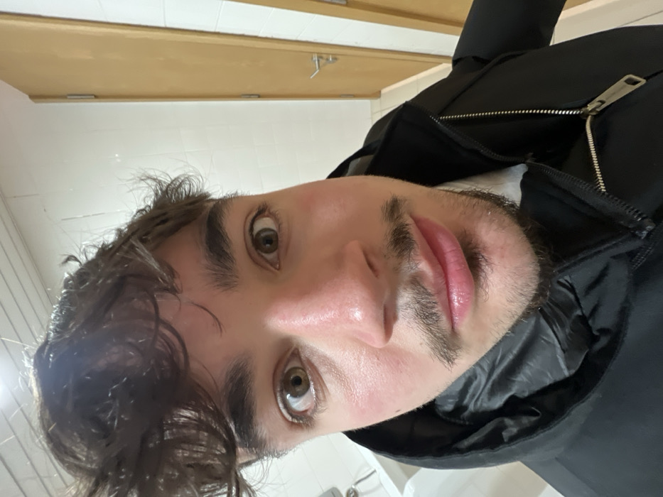 A selfie-style shot of a fair-skinned individual with hazel eyes, dark hair, and light facial hair is centered. The person is wearing a dark jacket with a partially visible zipper. A door, possibly in a bathroom, is in the background. The image appears to be taken at an angle, adding an unconventional perspective. The lighting seems to be fluorescent, contributing to the overall coolness of the scene.
