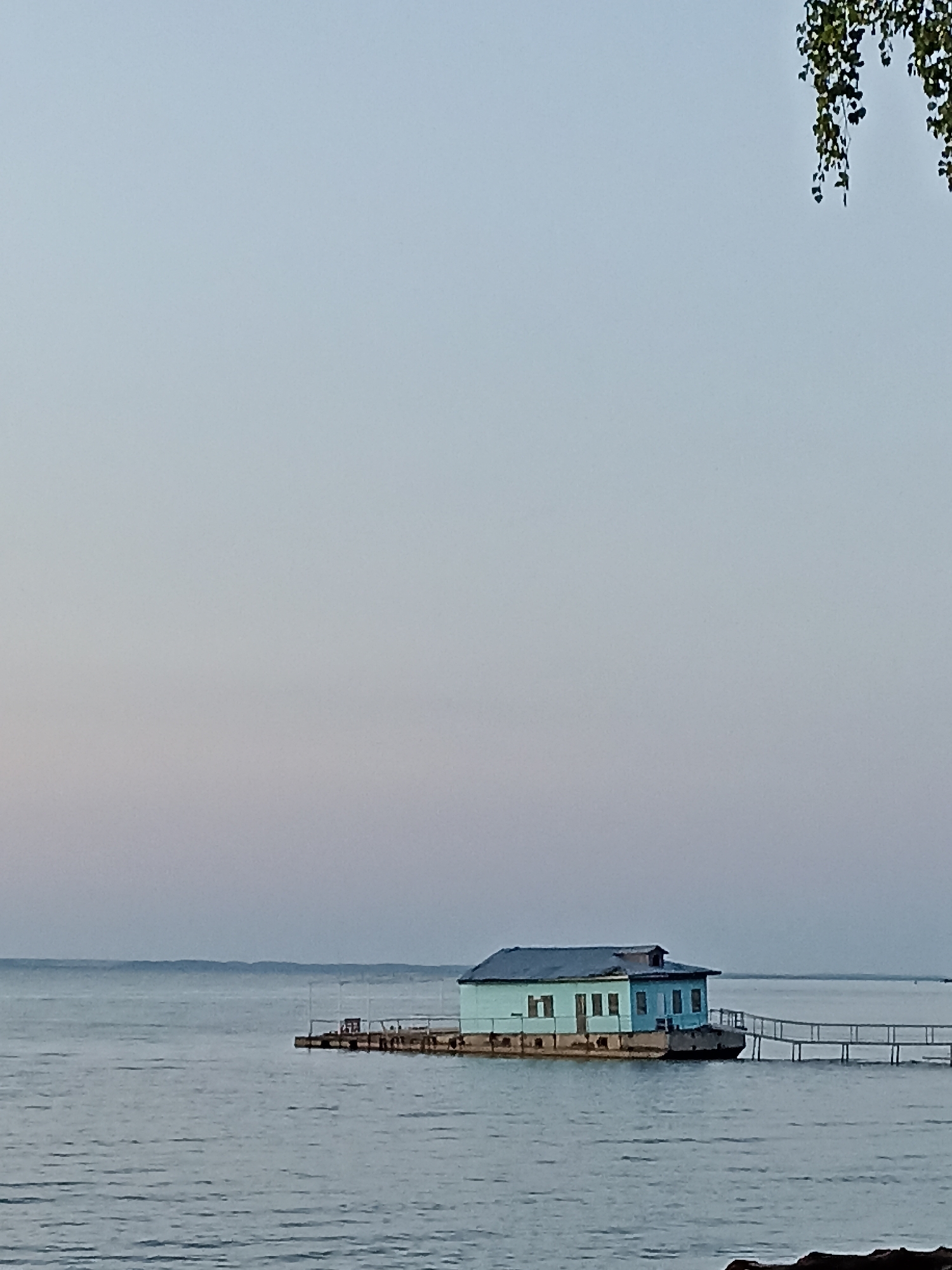 A pale blue, rectangular building sits on a concrete foundation floating on calm, gray water. The building has a dark roof and multiple windows. A wooden pier extends from the building to the right. The sky is a pale gradient of gray and blue, with tree branches hanging from the upper right corner. The scene appears to be a waterfront in Zavyalovo, Russia, possibly a boathouse or small dwelling. There are no people or animals visible in the image.