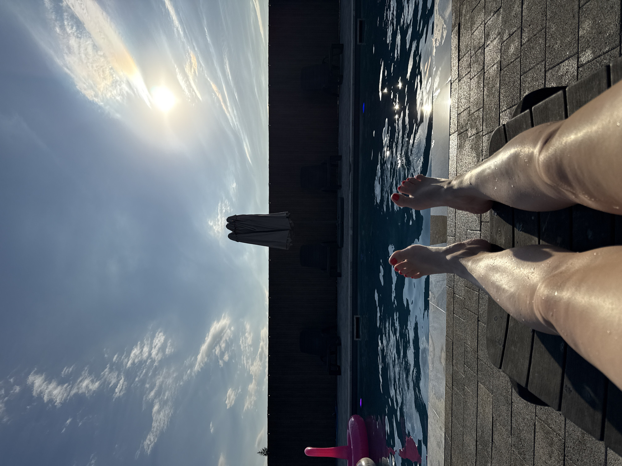 A first-person perspective shot shows a person's legs and feet relaxing on a dark-wood lounger. The person's legs are wet and glistening, their toenails painted red. The lounger is positioned on a stone patio overlooking a swimming pool. The pool is a deep blue, with ripples on the surface reflecting the bright sunlight. In the background, a dark wooden fence encloses the area, with two dark lounge chairs and folded umbrellas visible. The sky is a bright blue, with wispy white clouds and a sun halo visible. The overall scene suggests a peaceful summer day at a private residence. There is no identifiable information to place this scene in Pervorechenskoye, Russia.