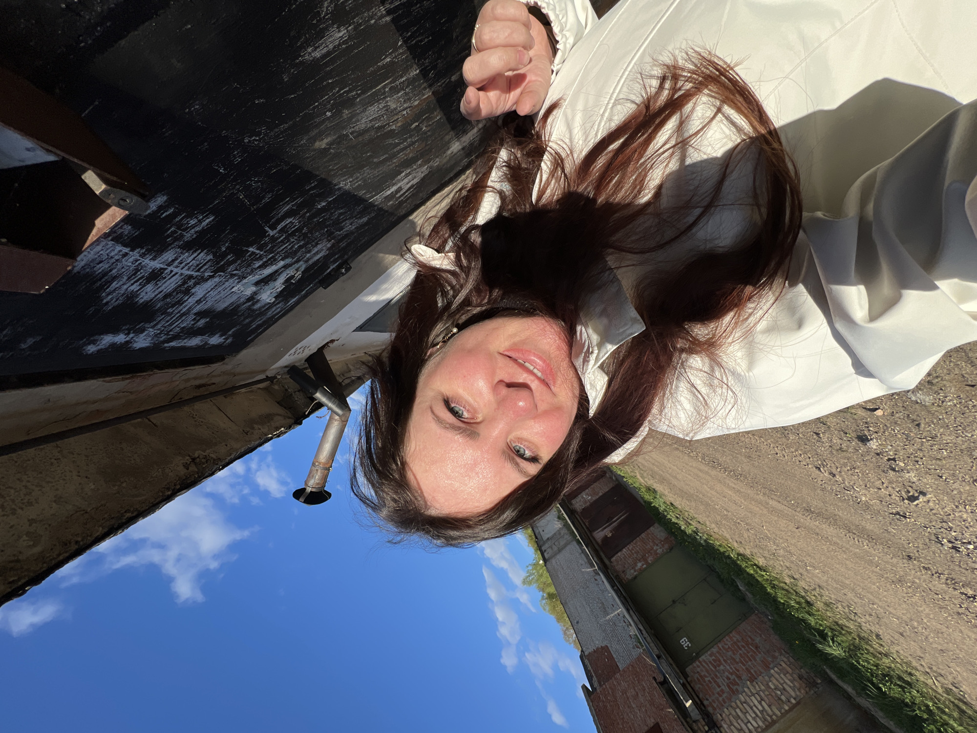 A woman with light skin and brown hair, wearing a white garment, is seen in an outdoor setting. She has blue eyes and a slightly upturned nose. The backdrop consists of a blue sky with scattered clouds, a weathered brick structure, a dirt area with green vegetation along the edge, and a dark, potentially wooden, surface overhead. A metallic pipe protrudes from the top left of the frame. A building or structure with what appears to be a metal door is partially visible.
I lack information to specify the exact location within the city of Noviy, Russia.