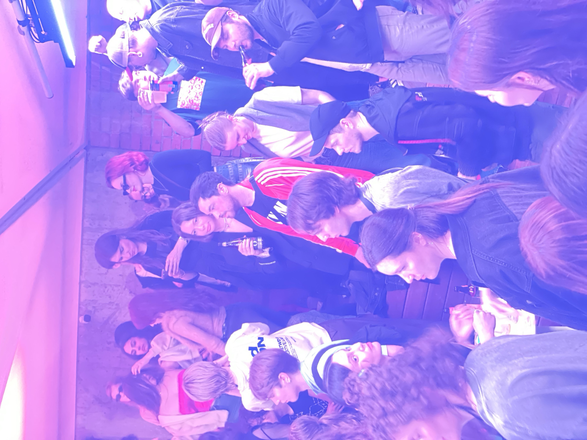 The image depicts a crowded indoor scene, likely a bar or club, illuminated by pink and blue lighting. A diverse group of young adults are present, many holding drinks or engaged in conversation. One person is smoking from a pipe. The environment suggests a social gathering with a relaxed atmosphere. The image is low resolution and grainy. The location is not specified in the image itself.