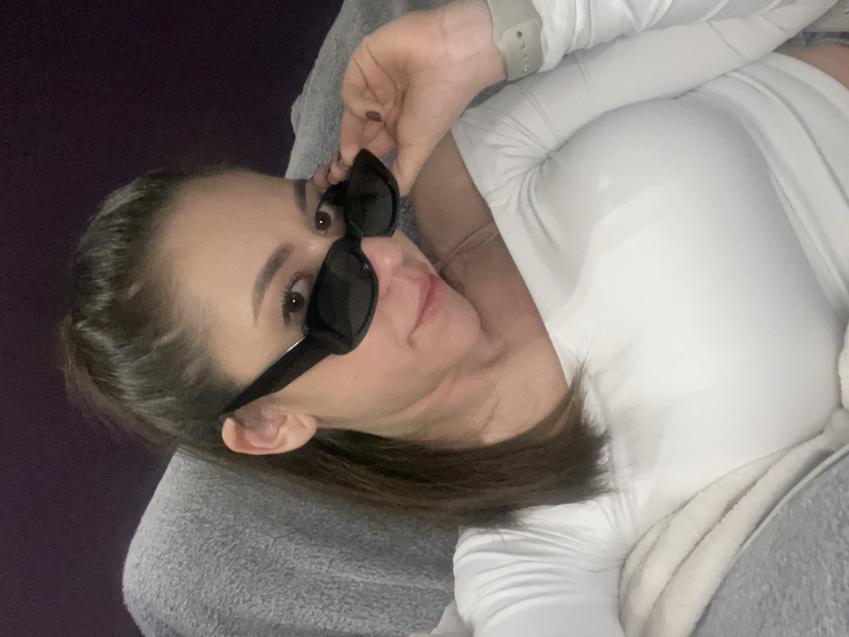 A female subject is depicted in an indoor, private setting in Sofia, Bulgaria. She has brown hair, styled in a high ponytail, and is wearing a white, off-the-shoulder, long-sleeved top. A thin, light-colored strap is visible on her right shoulder. The subject wears black, rectangular-framed sunglasses, which she is pulling down with her left hand, revealing her dark eyes. Her left hand's fingernails are painted dark, and a light-colored smartwatch is visible on her left wrist. She is looking directly forward, with lips slightly parted. The immediate environment includes a textured grey material, possibly a blanket or upholstery, on her left and lower side, and a plain dark purple wall in the background.