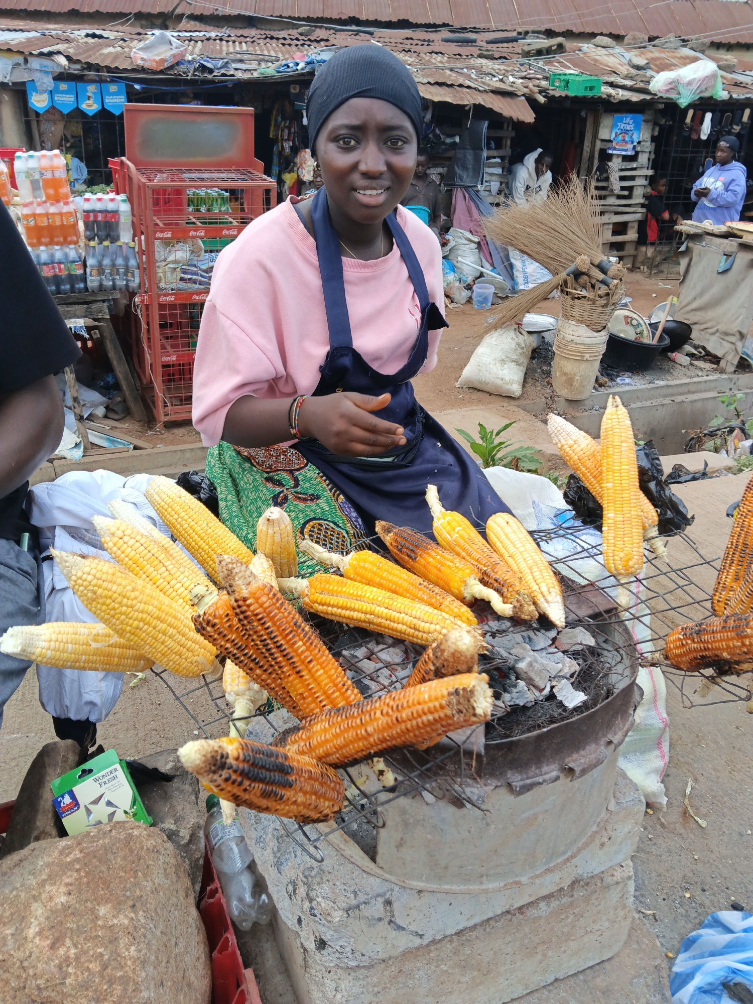 In Bukuru, Nigeria, a young woman wearing a black headscarf and a pink sweatshirt with a dark blue apron is tending to a charcoal grill laden with corn. The grill, made of metal, sits atop a makeshift structure of concrete blocks. Several ears of corn, some appearing freshly placed and others visibly charred from roasting, are arranged on the grill. Behind her, a market scene unfolds under a corrugated metal roof, with stalls selling various goods, including bottled drinks and other food items. To the left of the woman, a stack of bottled beverages is visible. The ground is dusty, typical of an outdoor market environment.