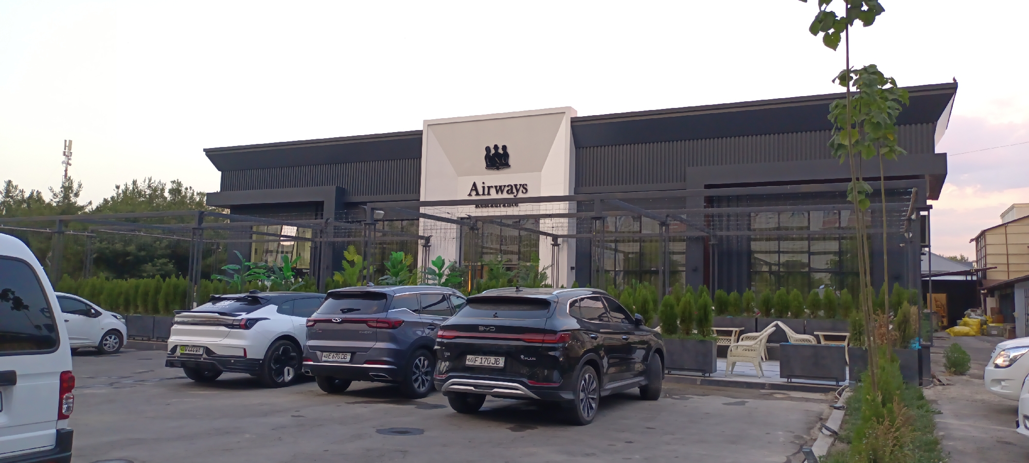 The image shows the exterior of a modern restaurant named "Airways" in Fergana City, Uzbekistan. The building has a sleek design with dark gray siding and a white facade featuring the restaurant's name and a logo of three figures. In front of the restaurant, there is a parking area with several cars, including a white van on the left, a white SUV, a dark gray SUV, and a black SUV on the right. The black SUV has a license plate that appears to be from Uzbekistan. The area is landscaped with rows of small evergreen trees and planters. The sky is overcast, suggesting it is either early morning or late afternoon. The overall scene depicts a commercial establishment with outdoor seating and a parking lot.