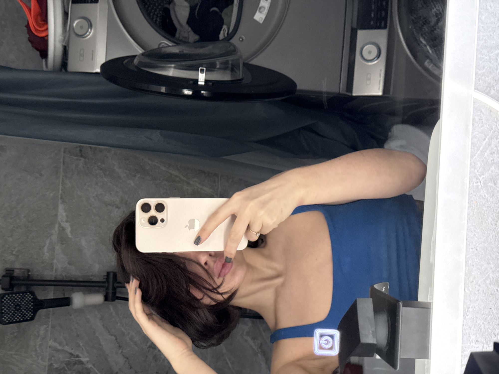 The image displays a mirror selfie taken by a woman in what appears to be a combined bathroom and laundry room, with the entire scene rotated ninety degrees counter-clockwise.
**Subjects:**
The primary subject is a woman, visible from the chest up, reflected in a large mirror. She has dark hair, which partially obscures her face. She is holding a light-colored iPhone with three camera lenses, using it to take the picture, concealing her nose and mouth. Her left hand (from her perspective) is visible near her neck/hair, and she wears a small ring on one finger. She is wearing a blue tank top.
In the background reflection, two silver-grey front-loading washing machines are visible, standing upright relative to the floor. To the left, a black showerhead and a black caddy or holder are mounted on a grey tiled wall.
**Activity or event taking place:**
The woman is engaged in taking a mirror selfie, holding her phone up to capture her reflection and the surrounding environment.
**Notable details:**
The scene is indoors, characterized by grey tiled walls and floor. One of the washing machines has a control panel showing the number "81". The woman's lips are visible, appearing to have some makeup, and her fingernails are painted with dark polish. A glowing, stylized power icon, resembling a digital overlay, is visible in the bottom right corner of the image. The ambient lighting suggests an indoor setting, without specific cues for the time of day.
**Visible text:**
The number "81" is clearly visible on the control panel of one of the washing machines. The Apple logo is visible on the back of the iPhone.