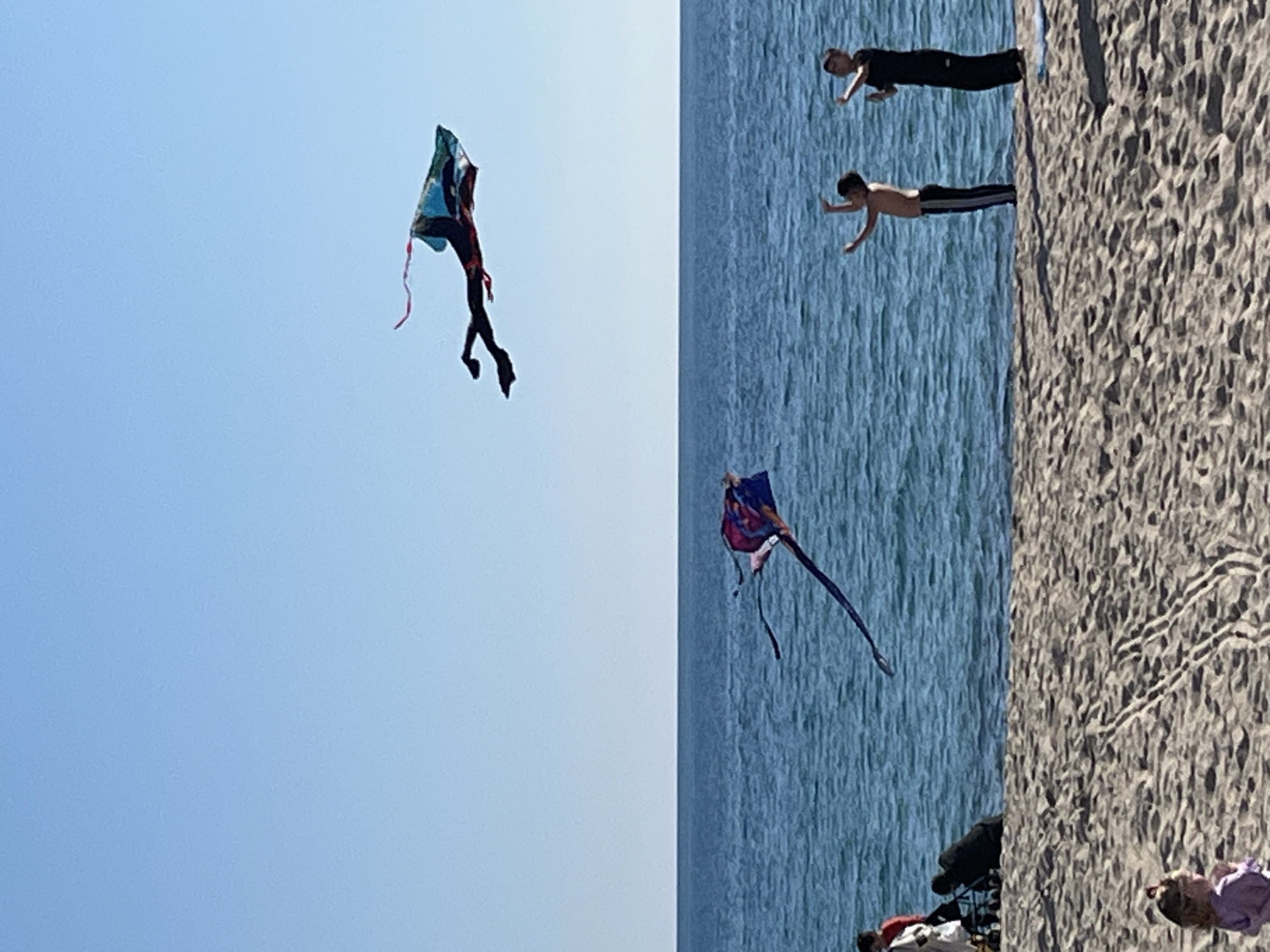 The image depicts a daytime scene on a wide, sandy beach in Alanya, Turkey, consistent with locations such as Cleopatra or Keykubat Beach, bordered by the Mediterranean Sea under a clear, light blue sky.
Two kites are actively flying. A large, dark-colored kite, predominantly blue, green, and black with red accents and trailing streamers, shaped like a stylized figure, is high in the upper right quadrant of the sky. A second, smaller, multicolored kite (purple, blue, black) with streamers is flying lower, positioned above the water line in the center-left.
Four individuals are visible on the beach. In the bottom left foreground, a young child wearing a light purple top is partially observed, facing away from the camera. On the mid-ground right, closer to the water, two individuals stand with arms raised, facing the sea. One is shirtless, wearing dark trousers with a white stripe. The second individual is fully clothed in dark attire, standing slightly behind the first. Their posture suggests active engagement with or observation of the kites. On the mid-ground left, a black stroller is situated on the sand. An individual in a red top and white trousers is partially visible next to the stroller, accompanied by another individual in dark clothing who is hunched over near the stroller. The sandy surface exhibits various footprints and linear patterns. The surface of the sea is mildly rippled.