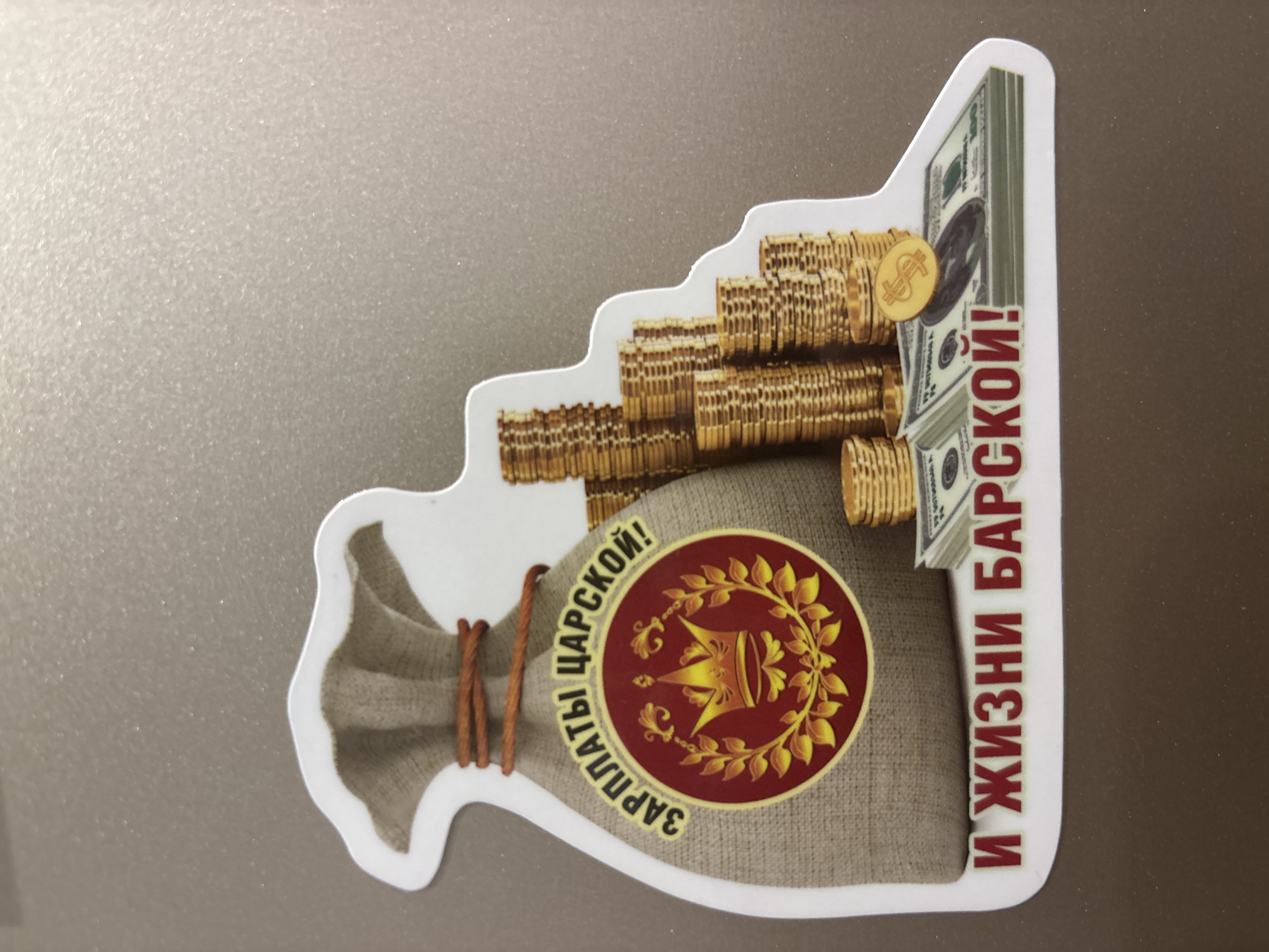 The image shows a sticker with a cartoon-like depiction of a bag, stacks of coins, and stacks of US dollar bills. The bag is light brown and tied with brown cords. It has a red and yellow emblem featuring a crown and laurel leaves, with the words "ЗАРПЛАТЫ ЦАРСКОЙ!" encircling it. The coins are gold, stacked in graduated levels like steps. A gold coin with a dollar sign rests on top of the stacks of US dollar bills. To the right of the money is the Russian text "И ЖИЗНИ БАРСКОЙ!". The background the sticker is on is a neutral gray. There is no indication of location of the city of Zagatuy, Russia within the image.