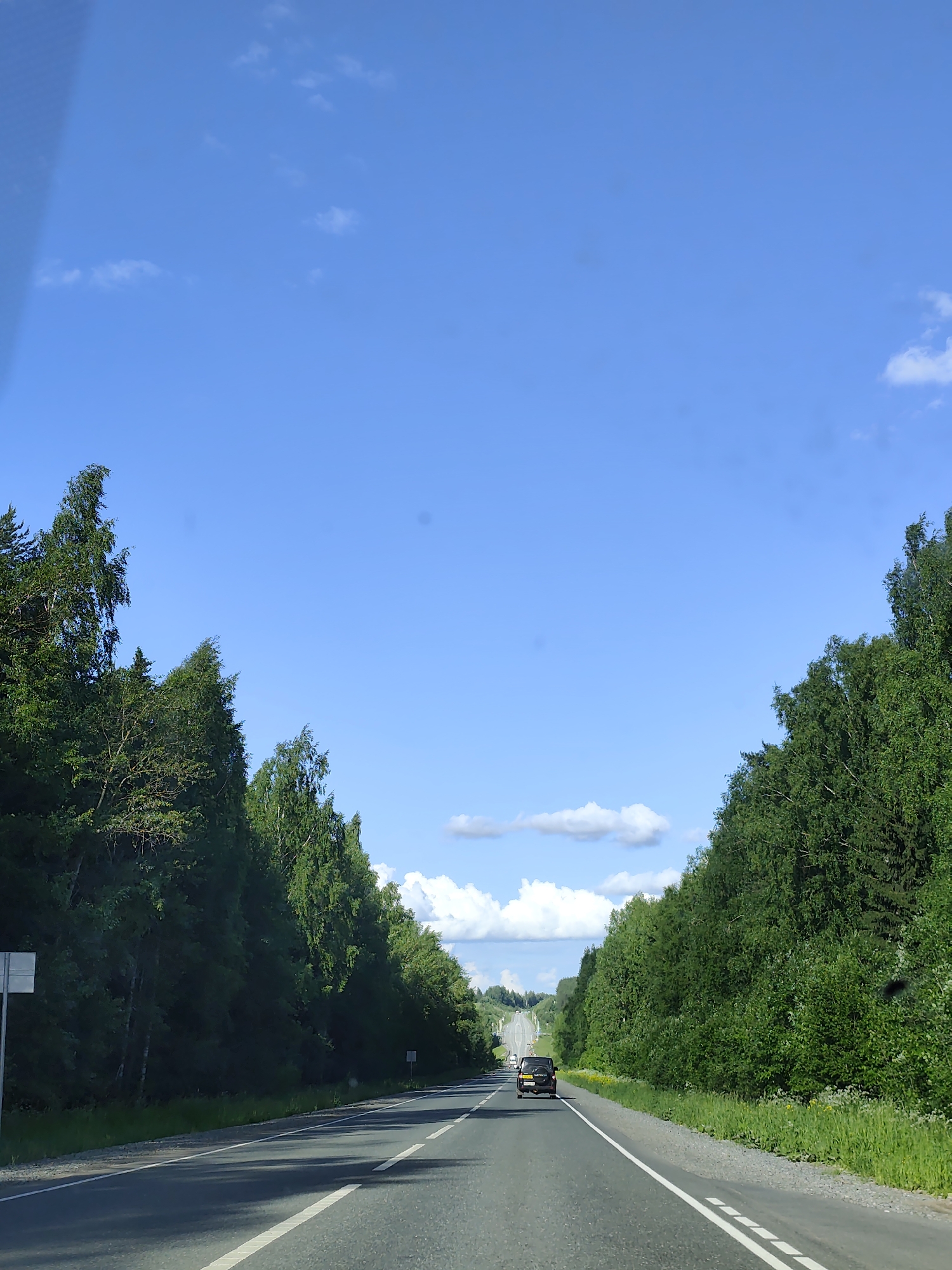 This is a vertical, eye-level, full shot of a paved road stretching into the distance, flanked by dense green trees on both sides. The sky above is a bright, clear blue with scattered white clouds. In the foreground, the road has white lane markings. A black SUV is visible driving away from the camera in the middle distance, on the right lane. The trees are lush and full, indicating a warm season. The overall scene depicts a rural or semi-rural setting, likely a highway or main road in Russia, possibly near the village of Bannoе, known for its natural beauty and recreational areas.