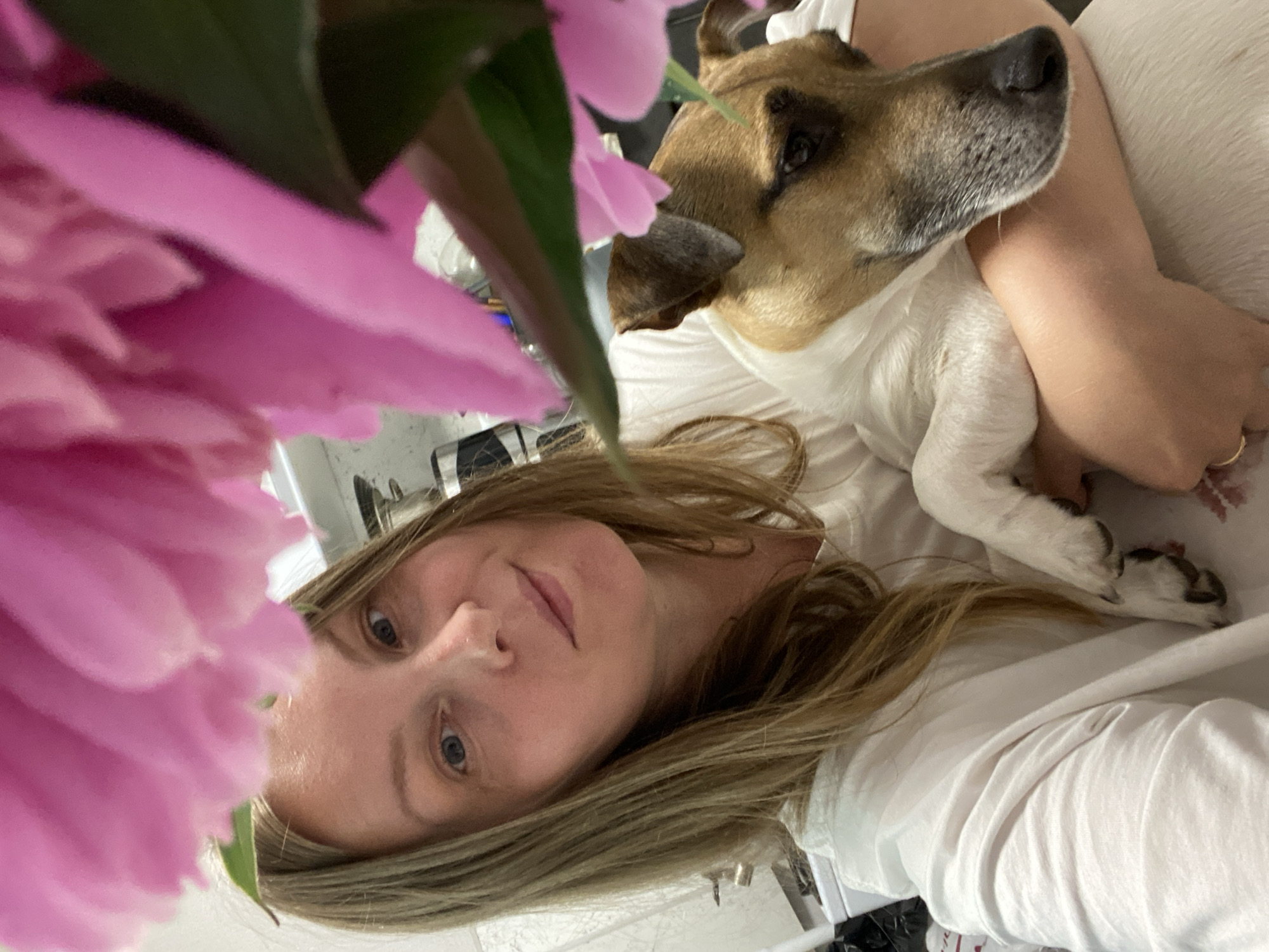 In Noviy, Russia, a woman with blonde hair smiles for a selfie, holding a small dog in her arms. Large pink flowers, possibly peonies, are in the foreground, partially obscuring the woman's face and the dog. The background is blurred, but appears to be an indoor setting with kitchen appliances visible. The woman is wearing a white t-shirt, and the dog has a tan and white coat. The overall mood is casual and affectionate.