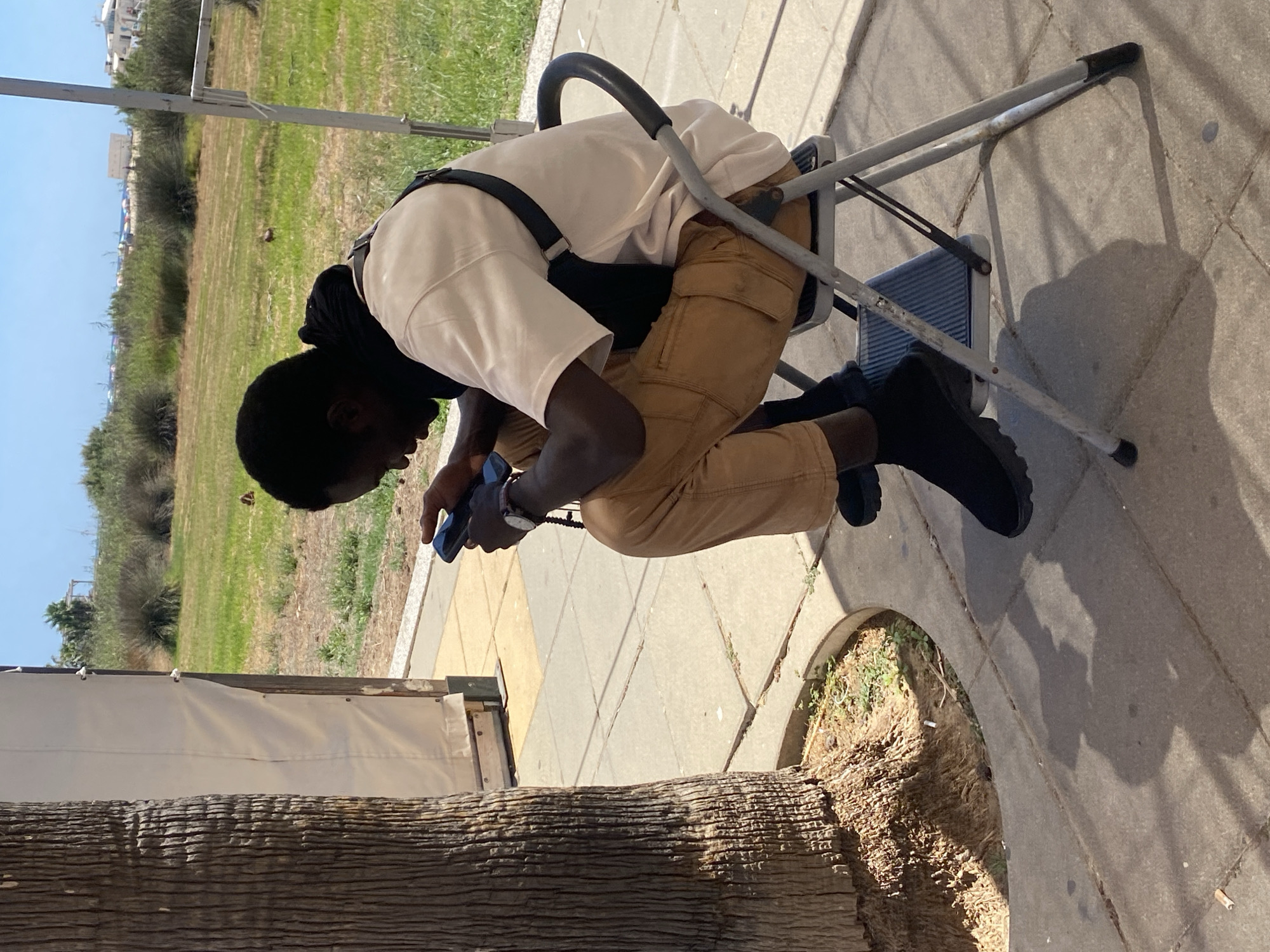 A dark-skinned man is seated on a folding metal chair, looking down at a black smartphone in his hands. He is wearing a light-colored t-shirt, tan pants, and black shoes. A dark bag strap is visible across his t-shirt. The man is sitting outdoors on a paved area with a textured brown tree trunk to his left. Beyond the paved area is a grassy field with some low bushes and trees in the background. The sky is clear and blue, suggesting a sunny day. The scene appears to be in a public outdoor space, possibly a park or recreational area, within the city of Conil De La Frontera, Spain.