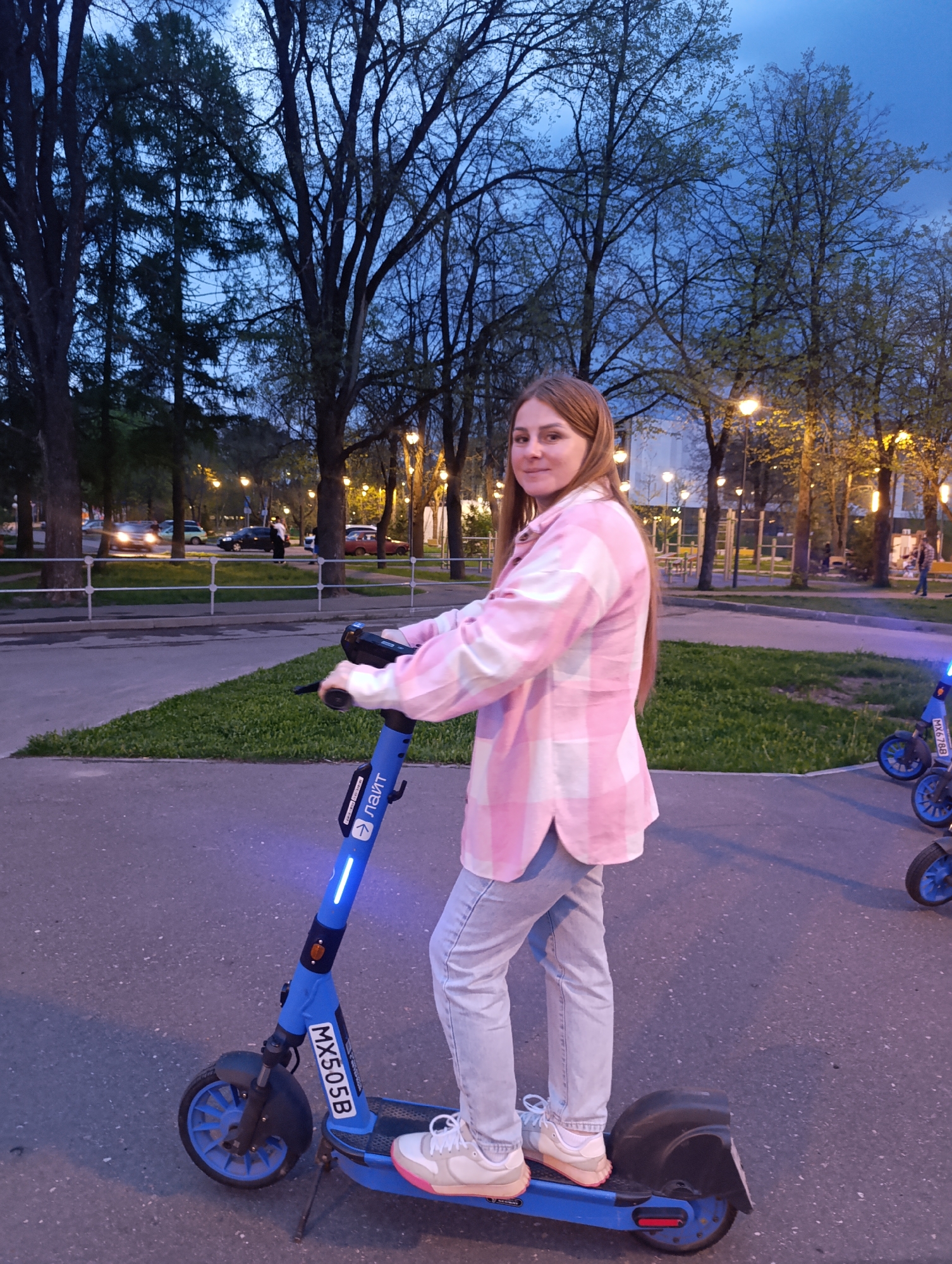 A person is riding a blue electric scooter on a paved area, likely a sidewalk or path, in the city of Unknown, Unknown. The person is wearing a pink and white plaid shirt, light blue jeans, and white sneakers with a pink accent. The scooter has the lettering "MX505B" on the front. The scooter has a blue glow illuminating the steering column. The background features trees, streetlights, grass patches, and other scooters parked nearby. It appears to be dusk or early evening, with a mix of natural and artificial light. Cars are seen further in the background.