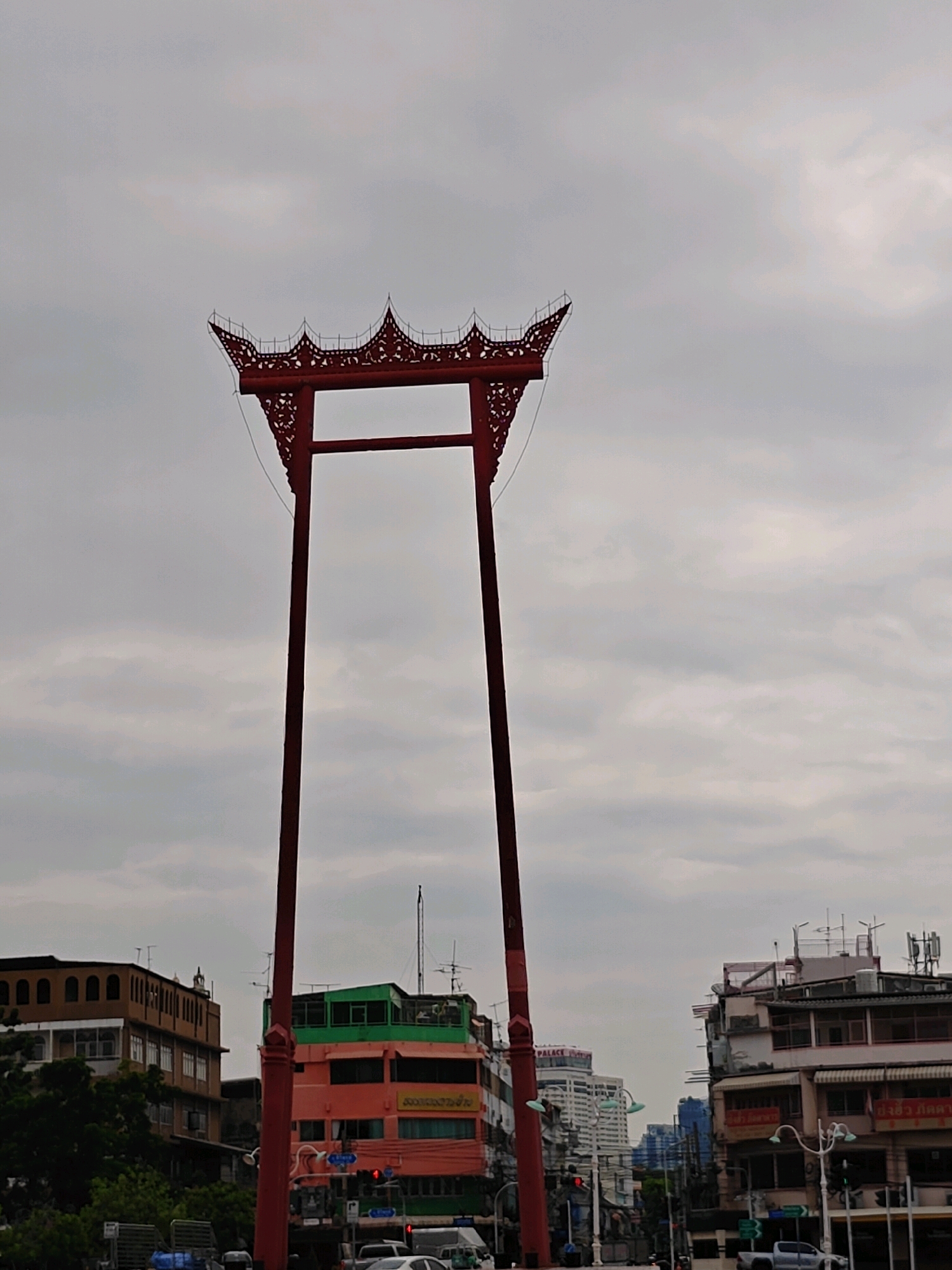 The image depicts the Giant Swing, a historical landmark in Bangkok, Thailand, located in the city's historic center near Wat Suthat. The swing, made of red painted wood and adorned with intricate carvings, dominates the upper center of the frame against a cloudy sky. Below the swing, a street scene unfolds with several multi-story buildings. To the left, a brown building with arched windows and a mosque-like dome is visible. In the center, a vibrant pink and orange building with green trim stands out, featuring signs in Thai. Further to the right, a building with balconies and shops is seen. The street is populated with traffic, including cars and trucks, and streetlights and traffic signals indicate an urban environment. The overall atmosphere is overcast, suggesting a subdued lighting condition.