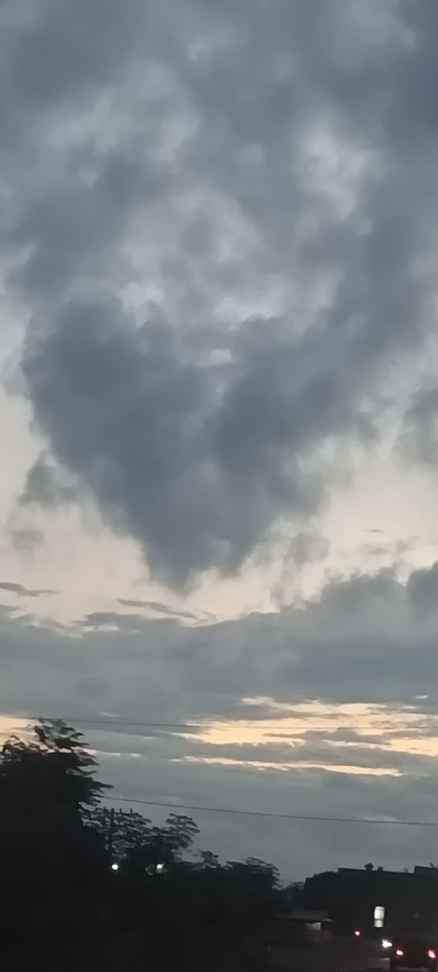The image presents a twilight sky, dominated by dense, grey, and overcast clouds. A distinct heart-shaped cloud formation is clearly visible in the upper-middle portion of the frame. Below the heavy cloud cover, a strip of lighter sky peeks through near the horizon, exhibiting soft transitions from pale yellow to light orange, suggesting either sunrise or sunset.
The lower section of the image features a dark, silhouetted landscape. On the left, several trees with dense foliage are visible, while on the right, dark shapes that appear to be buildings or structures extend towards the horizon. A thin, horizontal line, likely a power line, cuts across the middle ground. Several small, bright points of light, consistent with artificial illumination from streetlights or vehicles, are discernible within the darkened foreground.
The scene conveys a calm and somewhat muted atmosphere, with the soft, diffused lighting characteristic of low-light conditions. The prominent heart-shaped cloud adds a unique and emotional detail to the cloudy sky. No human figures or discernible text are present. The setting, located in Meunasahmee, Indonesia, appears to be an outdoor scene looking towards a distant settlement or road during the transitional period of day and night.