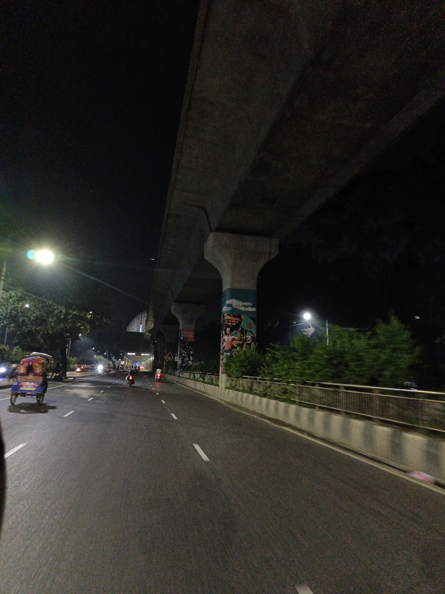 The image depicts a street scene at night in Dhaka, Bangladesh, likely under an elevated expressway. In the foreground, a blue and red auto-rickshaw is seen from the rear-left, moving forward. To the left of the road, a bright street lamp illuminates a large tree. The road is paved and has white lane markings. Several motorcycles are visible on the road, moving in the same direction as the auto-rickshaw.
The elevated expressway structure is prominent, supported by large concrete pillars. One pillar has colorful graffiti depicting people with flags. To the right of the road, there is a concrete barrier with a metal railing, and dense green foliage is visible beyond it. The background is dark, indicating nighttime. The overall atmosphere suggests a typical urban thoroughfare in Dhaka during the evening.