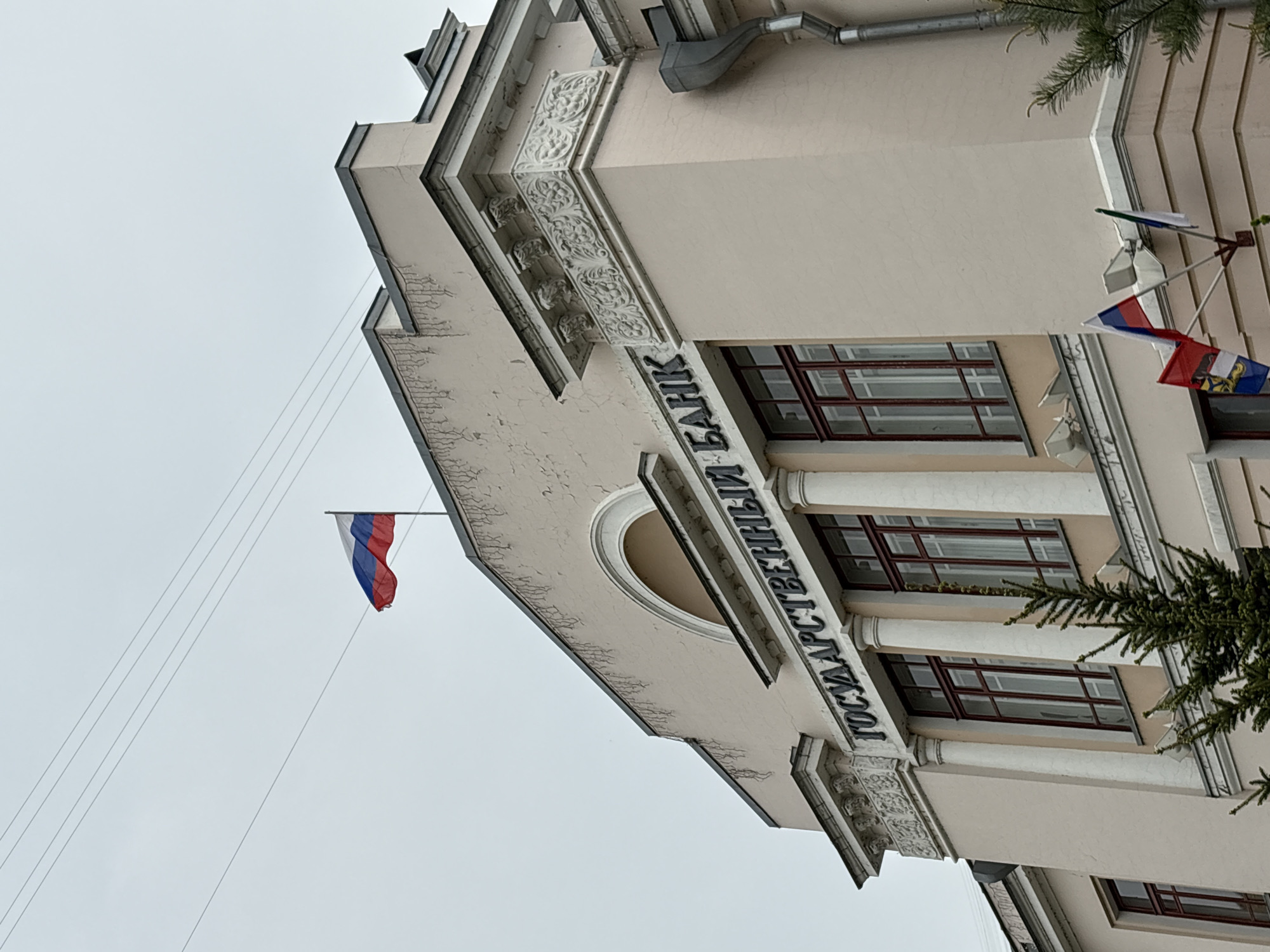 The image shows the facade of a building in Khabarovsk, Russia, identified by the inscription "ГОСУДАРСТВЕННЫЙ БАНК" (State Bank). The building features classical architectural elements, including columns, ornate moldings, and arched windows. Several flags are displayed on the building's exterior, including the Russian tricolor and another with an emblem. A Russian flag is seen on a pole extending from the building's roof. Power lines are visible in the foreground against a cloudy sky.