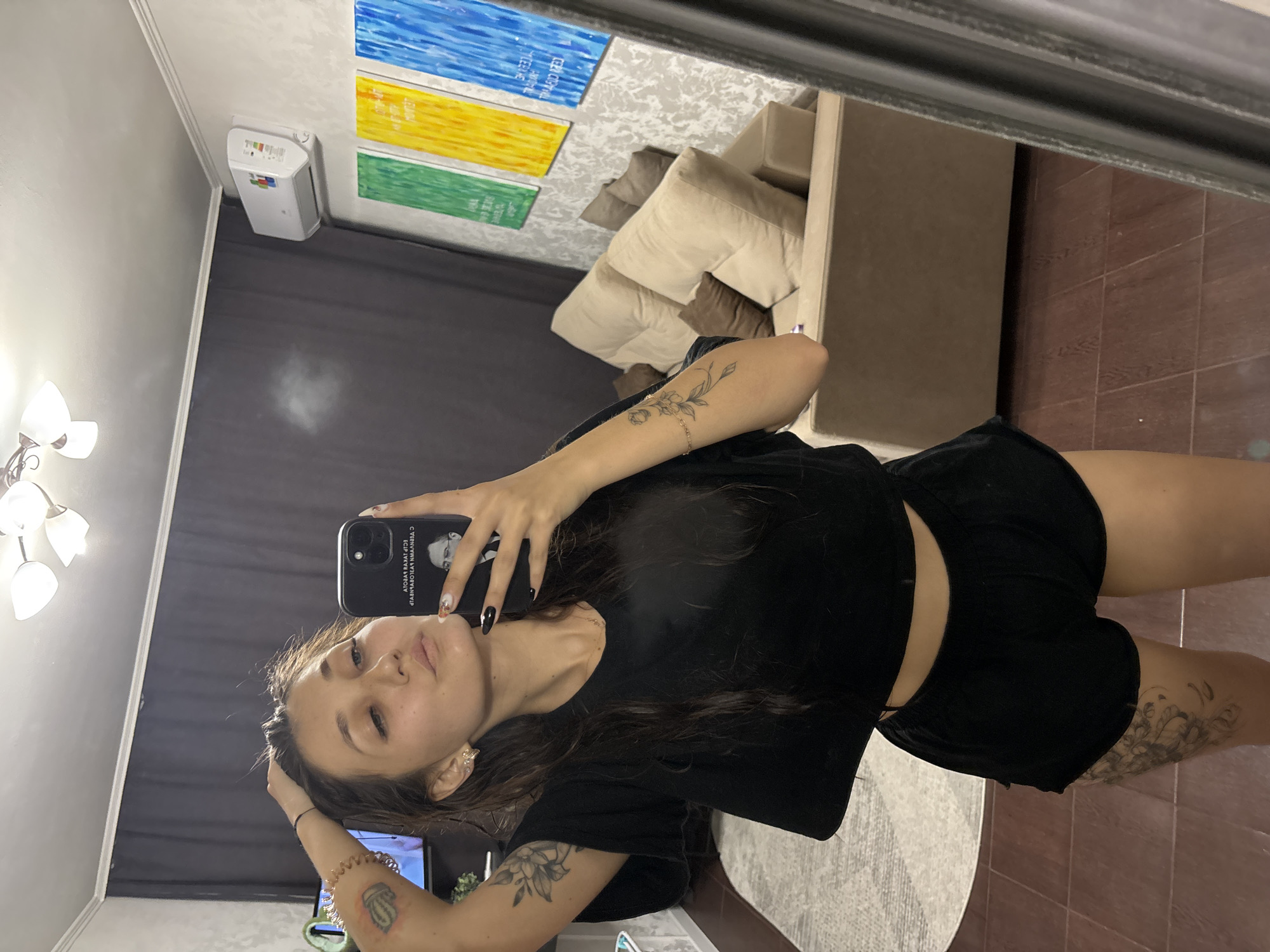 A young woman with dark hair and a black crop top and shorts is taking a mirror selfie in what appears to be a living room. She has tattoos on her arms and legs. In the background, a beige couch, colorful paintings on the wall, and an air conditioner unit are visible. The room has tile flooring and a patterned rug.