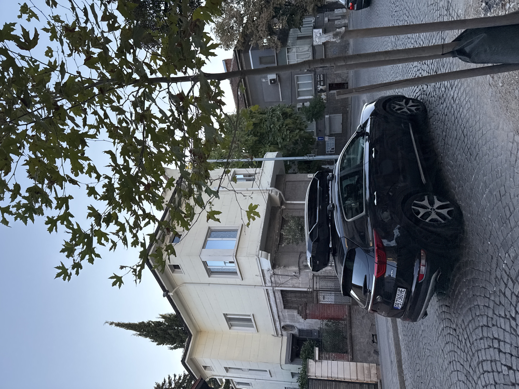 The image depicts a street scene in Plovdiv, Bulgaria, likely in an older neighborhood. A black Kia Sportage SUV, equipped with a roof box, is parked on a cobblestone street. The vehicle's license plate reads "PB 7360 HE". In the background, a pale yellow building with multiple windows is visible, with a tall, conical evergreen tree to its left. Adjacent to the yellow building is a darker, multi-story building with visible air conditioning units and windows. The street is paved with irregularly shaped cobblestones, and a mature tree with large, green leaves partially obscures the view of the buildings and the sky. The sky is overcast and pale. The overall atmosphere is calm and residential.