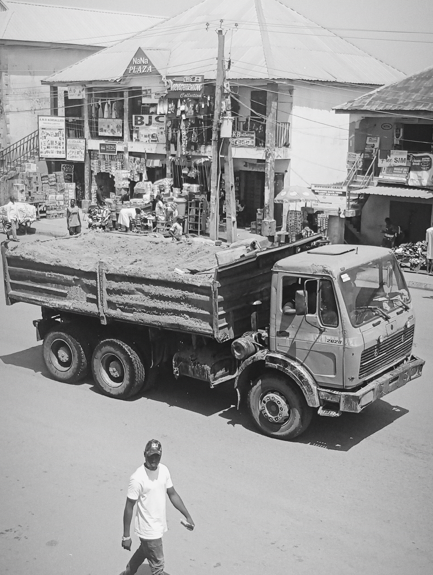 The black and white photograph captures a street scene in Bukuru, Nigeria, dominated by a large dump truck loaded with sand. The truck, a Mercedes-Benz model with the number 2628 visible on its front, is positioned in the foreground, its tires large and rugged. To the left of the truck, a man wearing a white t-shirt and a baseball cap walks away from the camera, holding a small object in his right hand.
In the background, a row of shops lines the street. Signs above the shops indicate various businesses, including "NaNa PLAZA," "Phenomenon Collection," "BJC," and "NEDU." Some shops display clothing and other goods. Several people are visible in and around the shops, some appearing to be customers and others shopkeepers. A woman stands near a stall, and other individuals are seen interacting in front of the businesses. The environment suggests a bustling urban commercial area, with overhead wires and a generally weathered appearance to the buildings. The overall lighting suggests it is daytime, with distinct shadows cast by the truck and overhead structures.