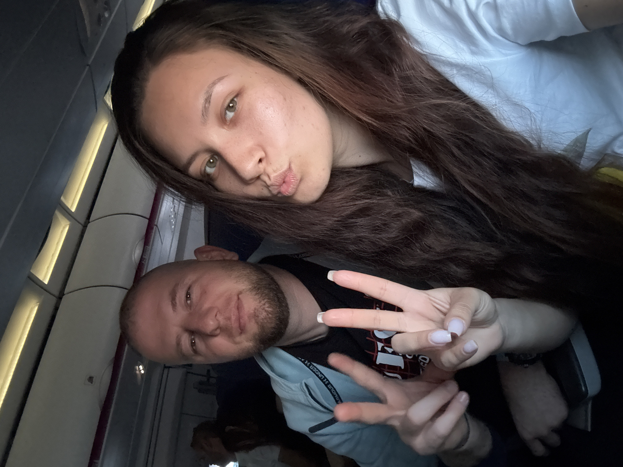 A medium shot captures a man and a woman posing for a selfie inside what appears to be an airplane cabin. The woman, positioned at the top of the frame, has long brown hair and fair skin. She is making a kissing face. The man, positioned below her, has a shaved head and a short beard. He is gesturing with both hands, holding up two fingers on each hand. He is wearing a dark T-shirt with a pattern on the front and a light blue jacket. The background shows typical airplane features, including overhead storage compartments and cabin windows. The scene takes place inside the airplane. There is nothing to connect this scene with the city of Rome, Italy.