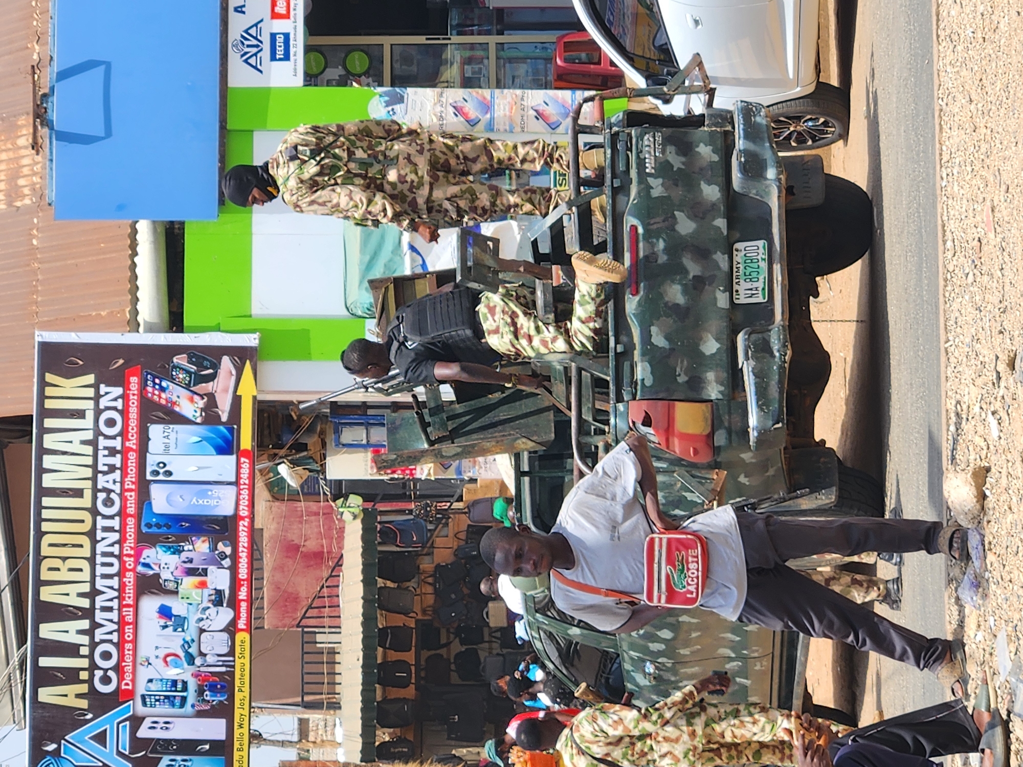 The image shows a street scene in an urban area, seemingly in Nigeria given the license plate information. There is a green and camouflage colored military pickup truck, with uniformed personnel in the back. One person is standing in front of the truck. The man is wearing a white shirt, black pants, and sandals, and has a Lacoste brand red shoulder bag. Behind the truck is a storefront with a large sign for "A.I.A ABDULMALIK COMMUNICATION," a business selling phones and accessories. The sign provides a phone number and address: 500 Bello Way Jos, Plateau State. Other people are present in the scene, but details about them are limited. The environment appears to be a commercial area with buildings and pedestrian traffic.