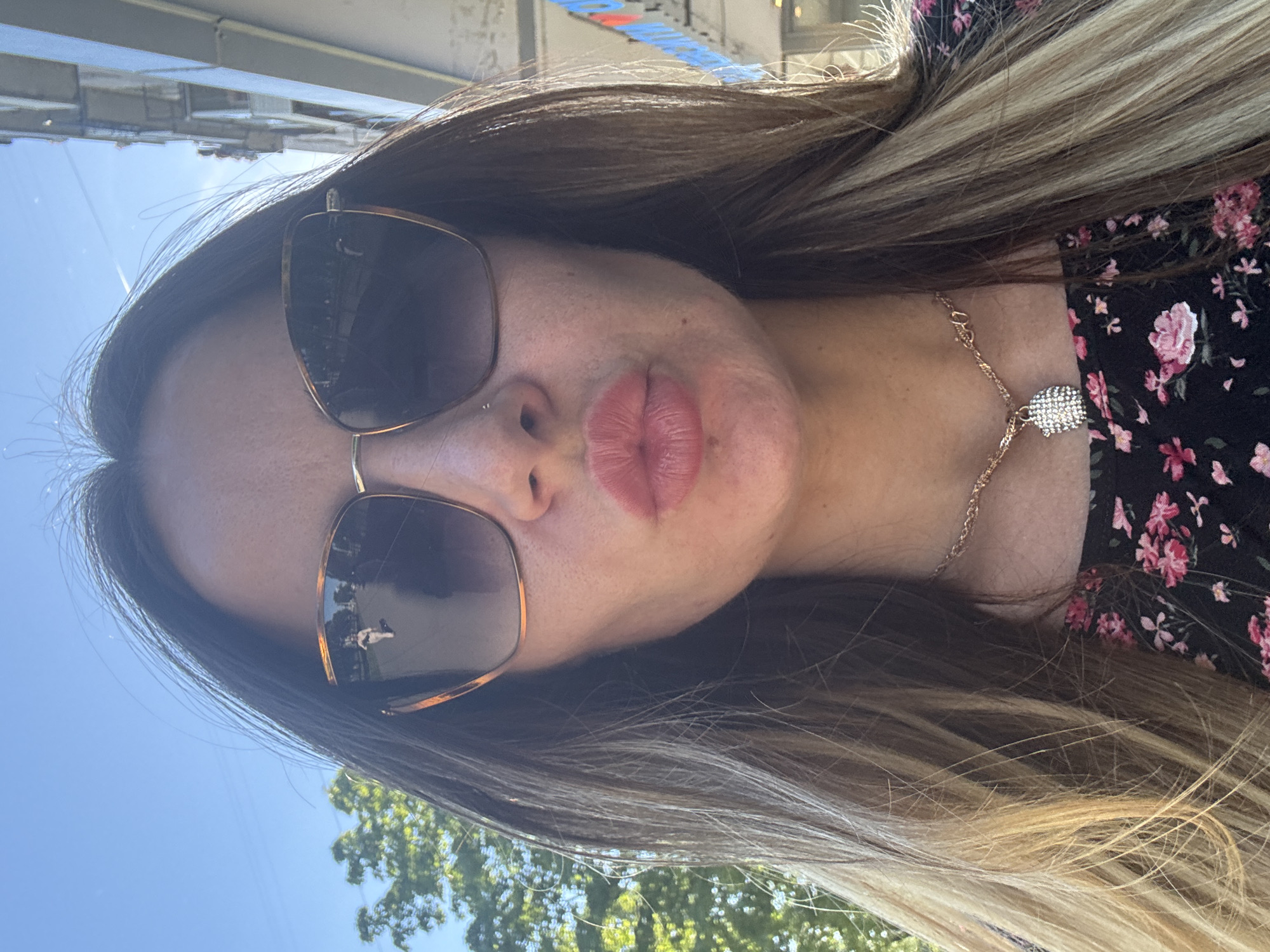 A woman with long, dark blonde-streaked hair wears oversized sunglasses and a floral print dress. She has a necklace with a sparkling pendant. Her lips are pursed, and a small nose stud is visible. The background shows a building and trees under a clear blue sky. The reflection in her sunglasses shows a person walking.