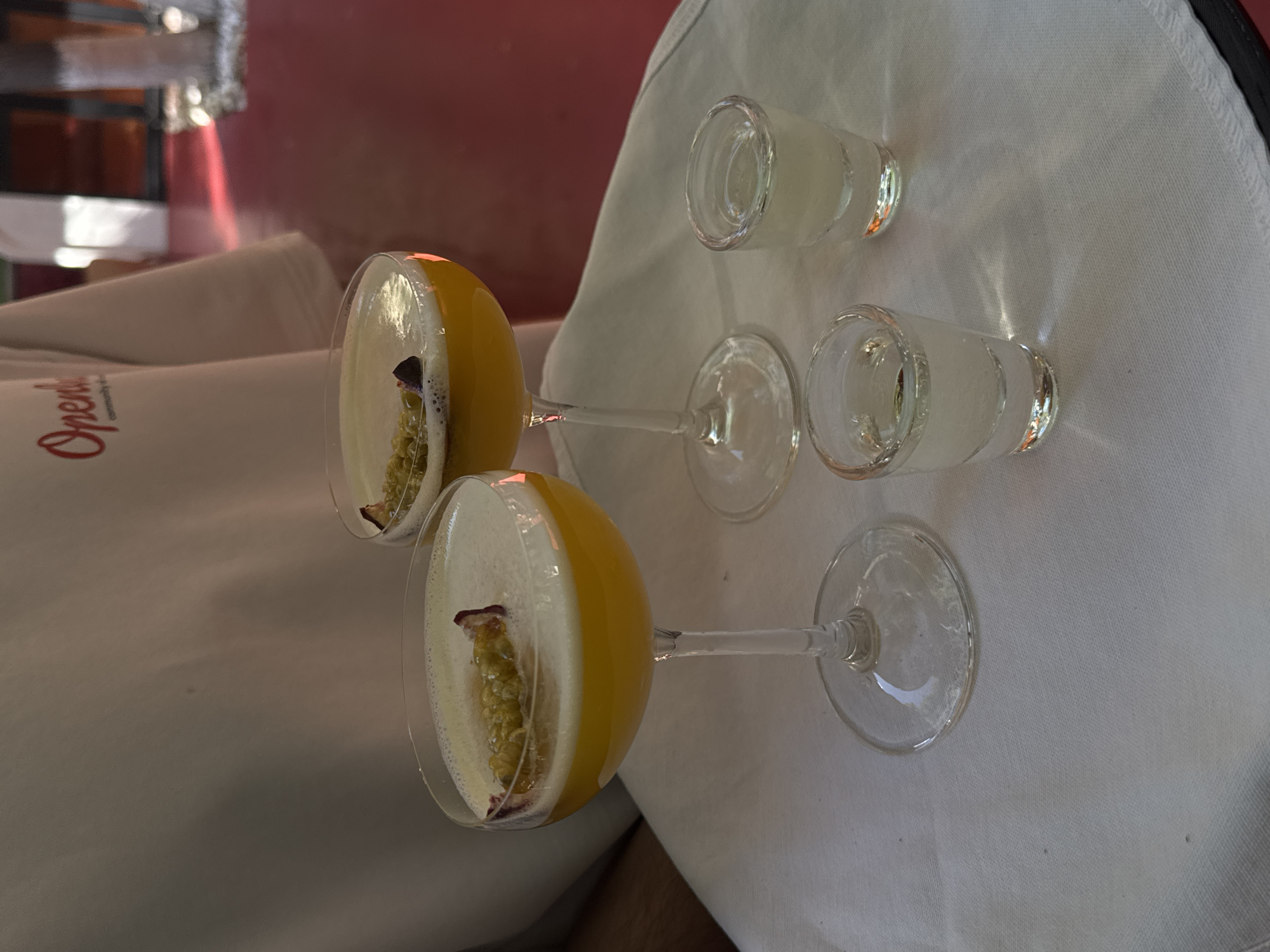 The image shows two champagne coupe cocktails topped with foam and garnished with passion fruit seeds and a purple petal. They are placed on a white cloth on a table. In the foreground, two shot glasses containing a clear liquid are also present. In the background, a person wearing a white t-shirt with red lettering that appears to say "Opendoor Community" is visible. The setting seems to be outdoors, possibly a cafe or restaurant, with a red floor and some greenery visible in the distance. The lighting suggests it is daytime. There are no specific elements that identify the location as Yerevan, Armenia.
