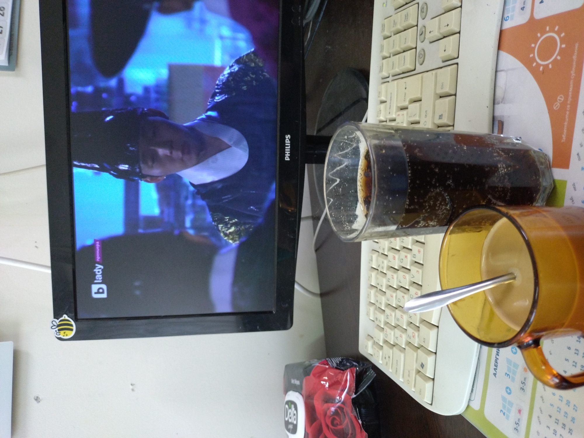 The image depicts a desk scene with a computer monitor displaying a historical drama or movie, a keyboard, a glass of dark beverage with bubbles, and a mug of coffee with a spoon. A package with a red rose design is partially visible to the left. The monitor shows a man in traditional attire and a high hat. The "PHILIPS" logo is visible on the monitor's bezel. A small bee sticker is affixed to the top left corner of the monitor. A calendar, possibly with Bulgarian text such as "АЛЕРГИЯ," is visible beneath the mug and keyboard. The overall setting appears to be an indoor workspace. There are no discernible elements that would pinpoint the location to Stara Zagora, Bulgaria.