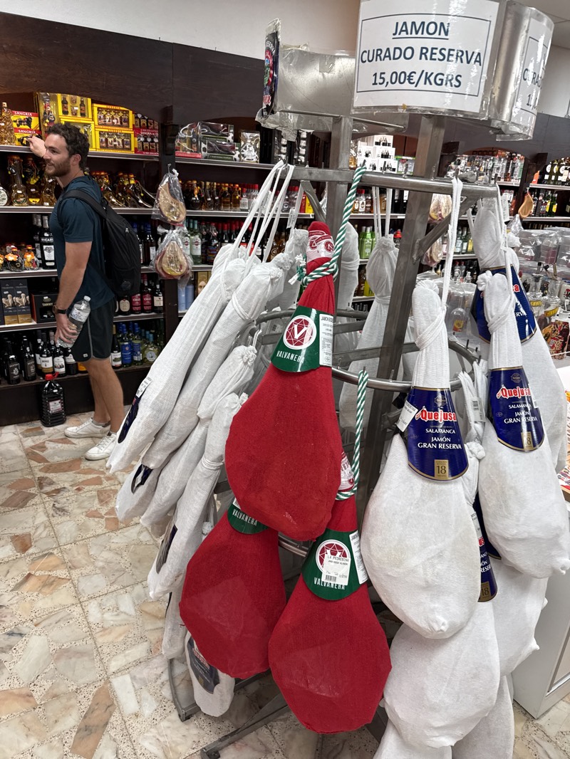 The image shows a grocery store in Fuentes de Oñoro, Spain. A fair-skinned male with dark hair and a dark shirt, shorts, and a black backpack is visible on the left of the frame. He is looking at items on a shelf containing alcohol and other goods. In the foreground is a display stand of cured ham legs, each individually wrapped in white or red netting. A sign above the stand advertises "JAMON CURADO RESERVA 15,00€/KGRS." Brands like Valvanera and Quejusa are visible on the hams. The store's tiled floor and fully stocked shelves of various products, including alcohol and packaged goods, are evident in the background.