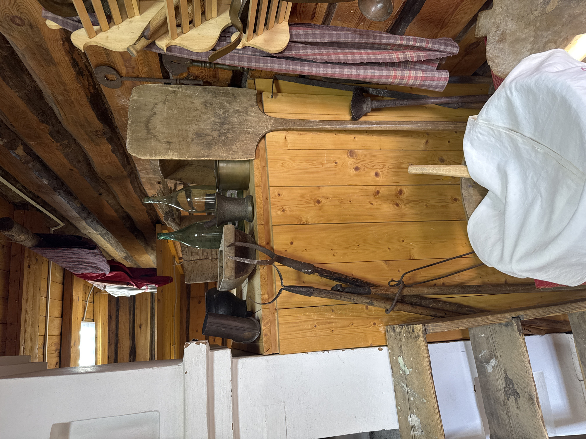 The image depicts an interior space with a variety of objects suspended from the ceiling or attached to the walls. The walls appear to be constructed of unfinished wood planks, while the ceiling consists of rough-hewn beams. Dominant objects include wooden cooking utensils, such as paddles and spatulas, along with metal tools like tongs and rakes. Several glass bottles are also visible, attached to the wall. A large white cloth object is present, possibly a sack or covering. A wooden ladder leans against the wall on the right side of the image. The scene appears to be a cluttered storage area or a display of historical artifacts. There are no people visible in the image. The location is specified as Votkinsk, Russia, indicating a possible historical context related to the region's cultural or industrial heritage.
