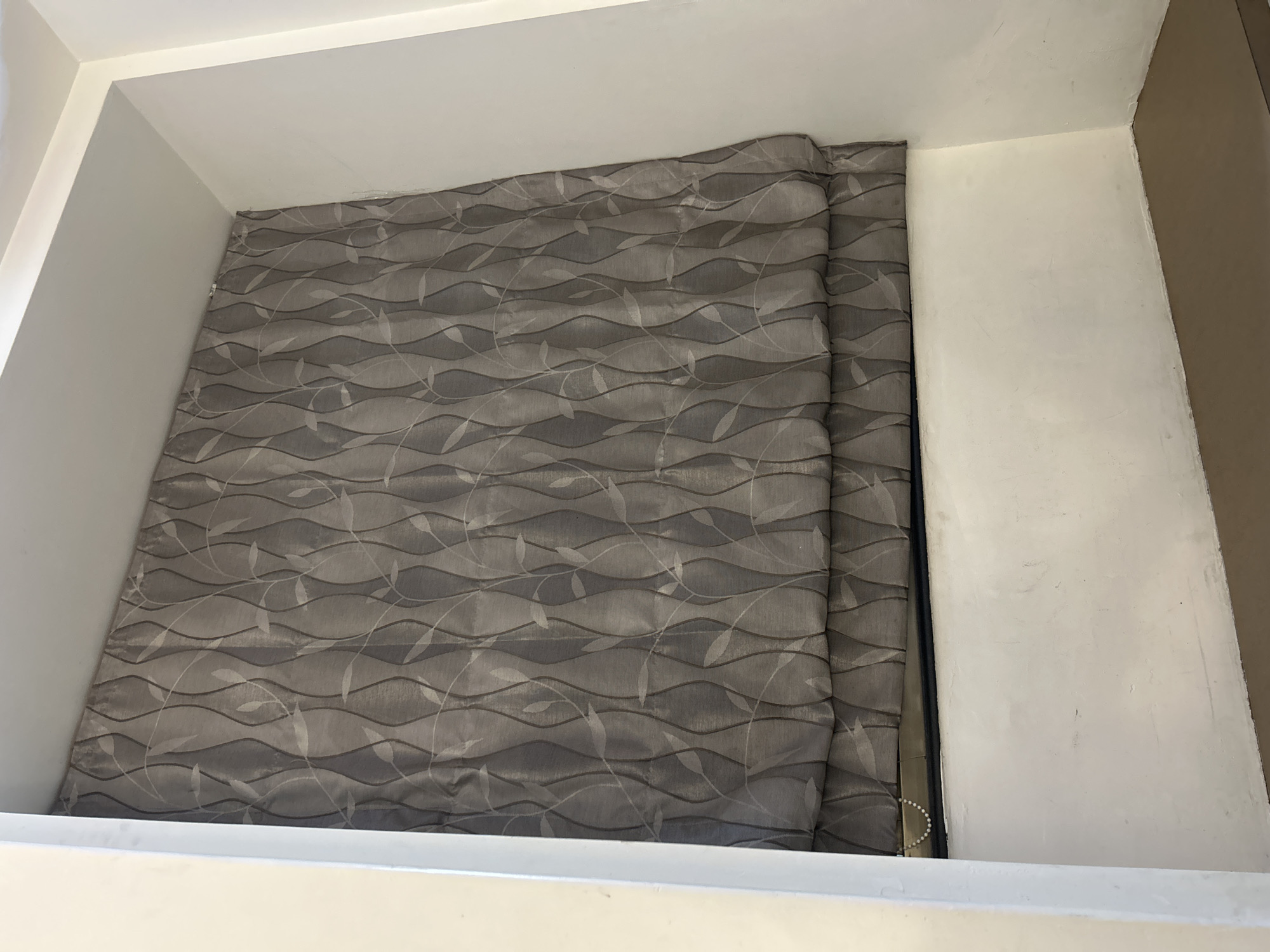 The image shows a gray roman blind with a leaf and vine pattern drawn across a window recess. The blind is partially drawn, creating folds at the bottom. A string of white beads hangs from the left side of the blind. The window recess is painted white, and the surface below the window is a brown ledge. The background wall is also white. The scene appears to be indoors, likely a domestic setting. There are no people or animals visible in the image. The location is not specified in the image.