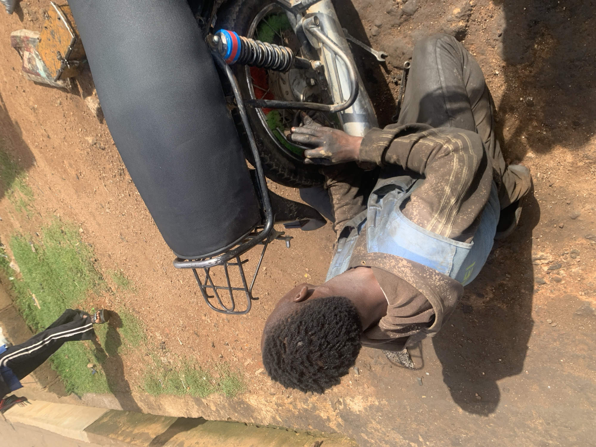 In Gura Top, Nigeria, a person is seen working on a motorcycle. They are wearing a brown hoodie and a blue vest, with their hands covered in grease. The motorcycle is dark and has a black seat. The ground is a mix of dirt and gravel. In the background, there is a small patch of grass and a concrete structure. A person's legs, wearing black pants with white stripes and sandals, are visible in the upper left corner of the frame, suggesting another individual is present or has recently been in the vicinity. Tools, including a wrench, are scattered on the ground near the motorcycle. The scene appears to be an outdoor repair area.
