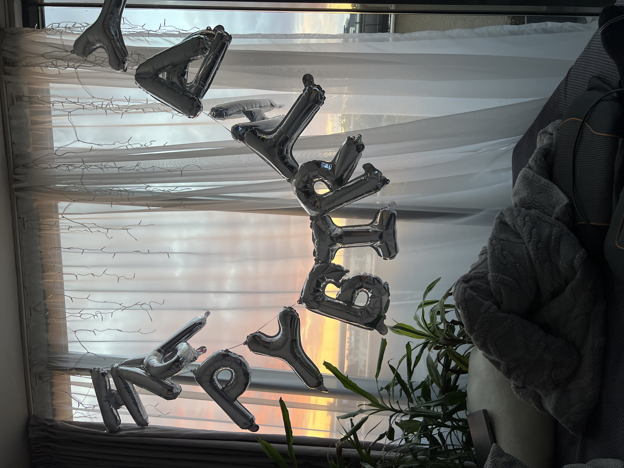 The image shows silver helium balloons spelling "Happy Birthday" hanging in front of a window with a sunset view. The balloons are attached to a string of fairy lights draped across the window frame. A potted plant sits on a surface near the window. To the right, part of a grey textured blanket or throw is visible. Outside the window, the sunset sky displays hues of orange and yellow. The location appears to be indoors. The city is not discernible from the visual evidence.