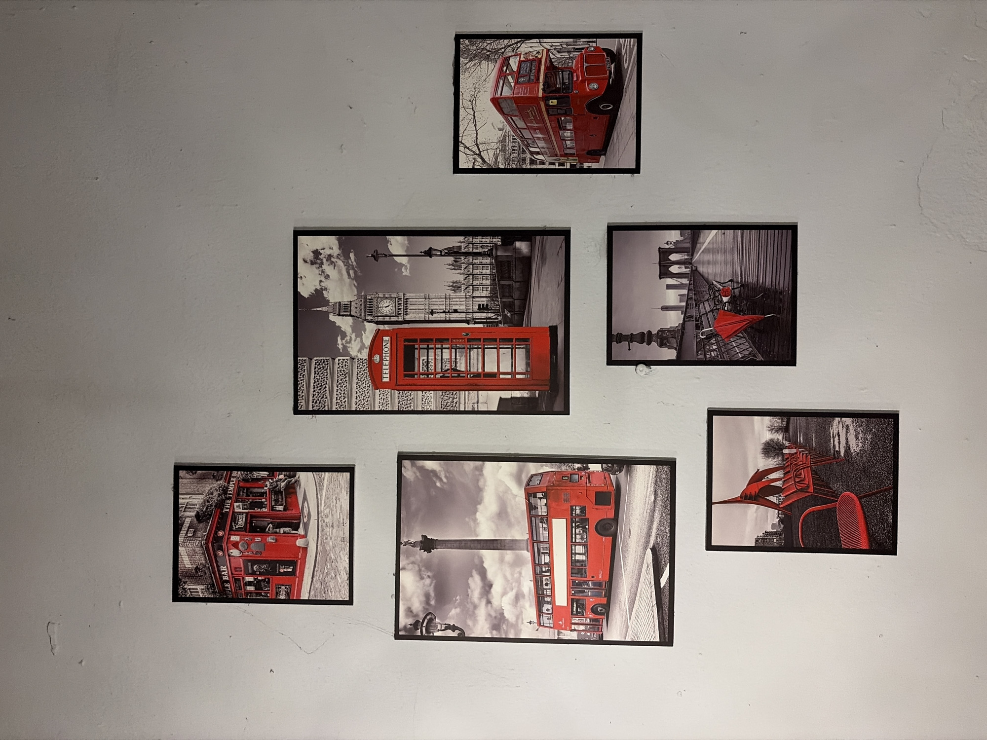 A collage of six framed photographs is displayed on a textured white wall. The photographs depict scenes from London, England, featuring iconic red double-decker buses, a red telephone box, and well-known landmarks like the Big Ben clock tower and Nelson's Column. The arrangement of the photos is asymmetrical, with a larger vertical photo of a red bus and Nelson's Column on the left, a vertical photo of a red telephone box and Big Ben on the right, and smaller photos distributed around them. The wall itself shows signs of wear, with visible cracks and unevenness. The lighting is even, suggesting an indoor setting. There are no people or animals visible in any of the photographs. The scene does not provide any specific details that would place it in Đà Nẵng, Vietnam; rather, it distinctly depicts elements of London.