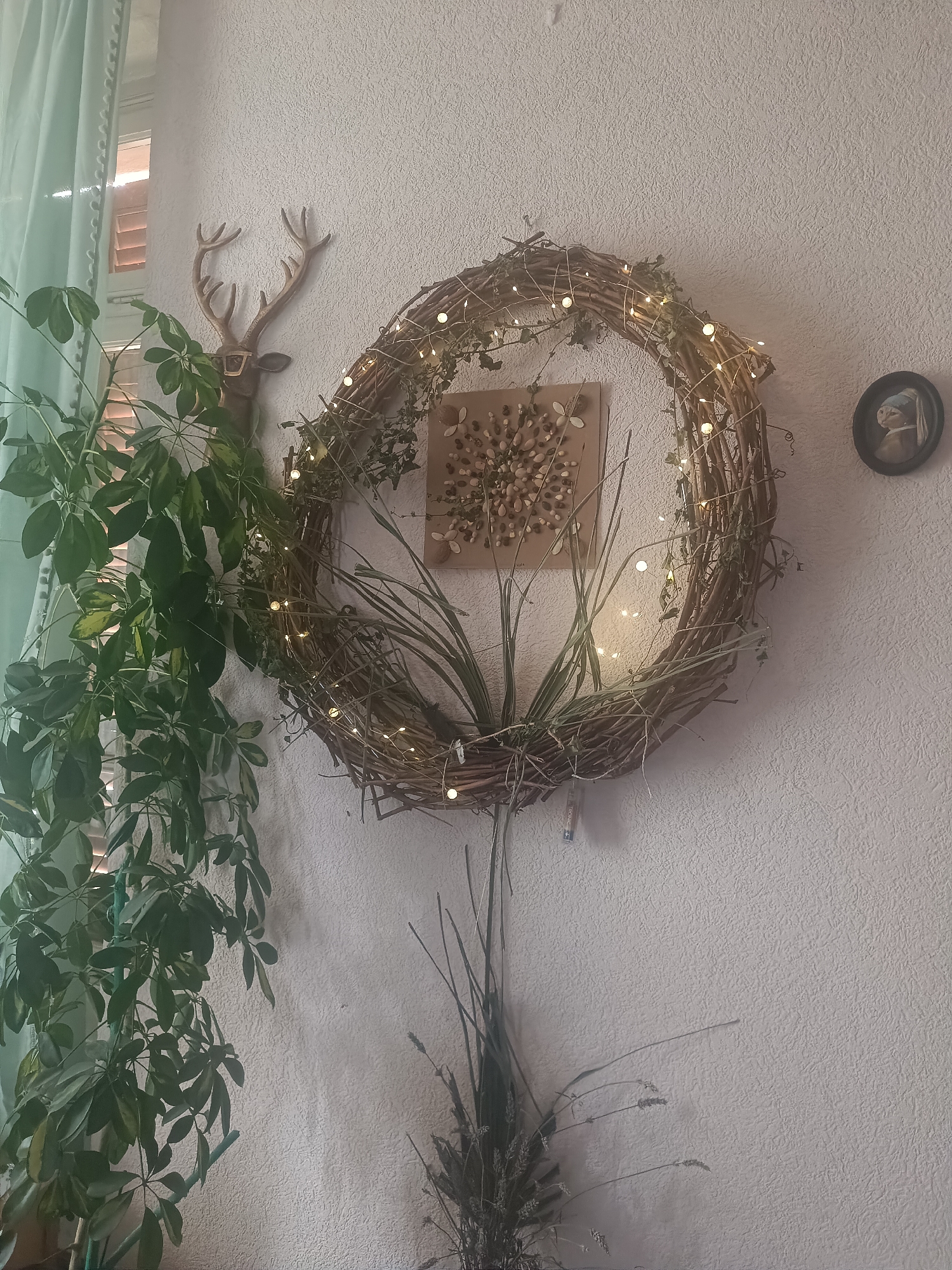 A decorative wall hanging featuring a woven wreath adorned with fairy lights and dried greenery is centered on a textured white wall. To the left of the wreath, a faux deer head with antlers and glasses is mounted on the wall. A large, leafy green plant cascades down from the upper left corner of the frame. Within the wreath, a square piece of cardboard displays a mosaic of small stones and seeds. To the right of the wreath, a small, circular framed image, resembling a miniature painting of "Girl with a Pearl Earring," is hung on the wall. Below the wreath, a bundle of dried grasses and flowers hangs. The scene appears to be an indoor decoration. The exact location within the city of Serravalle, Switzerland is not discernible from the image.