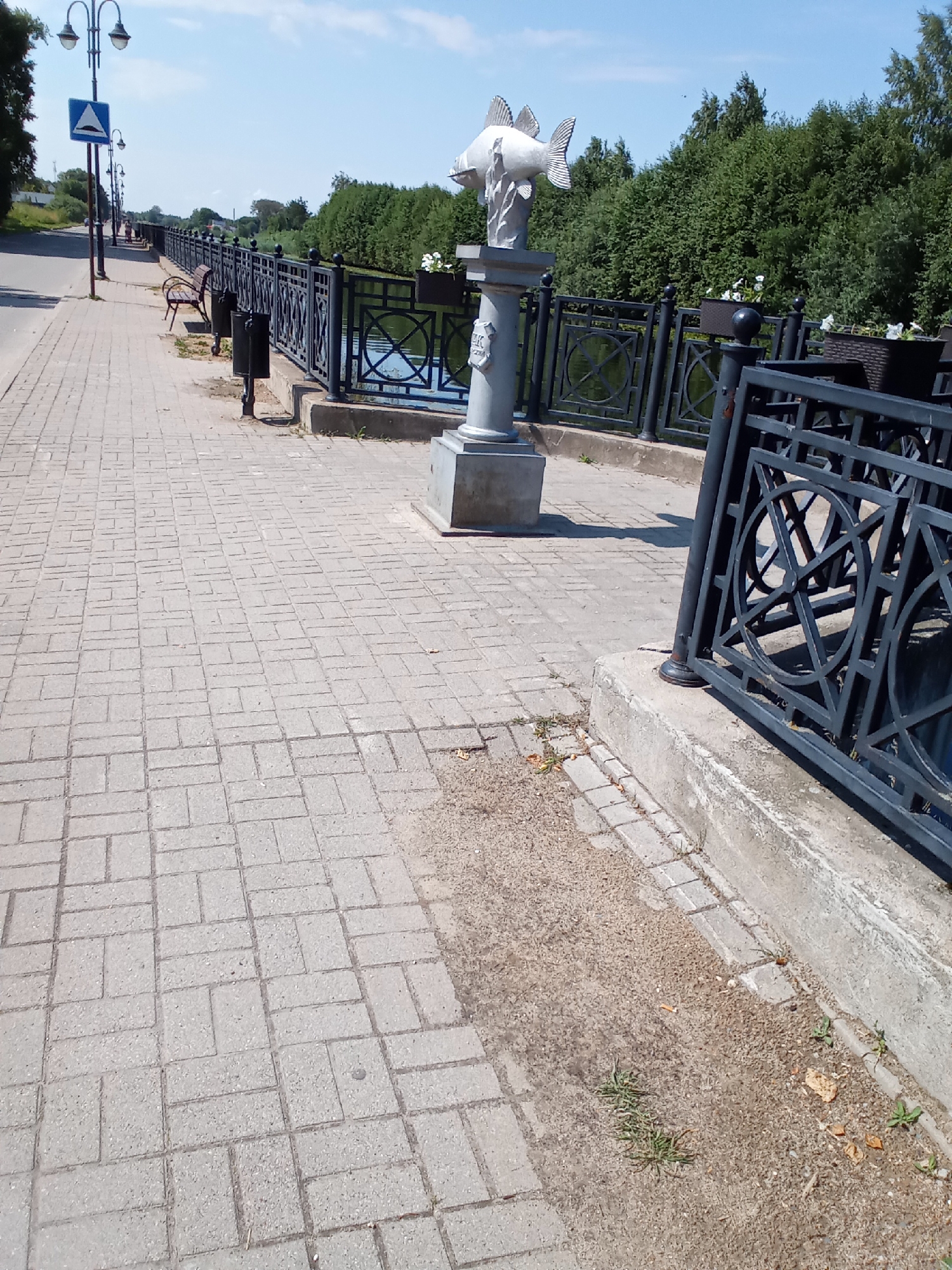 The image depicts a sunny day along a paved walkway next to a body of water. A decorative black metal railing with circular motifs runs along the edge of the walkway, separating it from the water. A stone sculpture of a fish sits atop a pedestal near the railing. The walkway is made of light-colored interlocking pavers. In the background, lush green trees line the opposite bank of the water. Further down the walkway, street lamps and benches are visible, as is a blue traffic sign indicating a speed bump. The scene is set in the city of Belozersk, Russia.