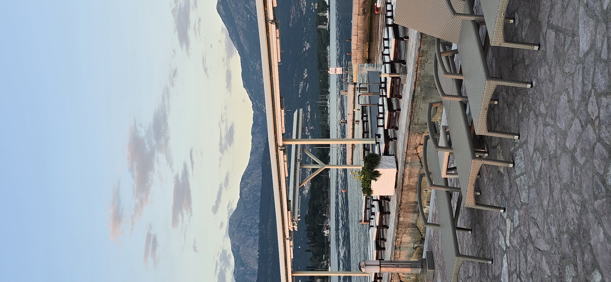 The image depicts a waterfront scene in Bogišići, Montenegro, at what appears to be an outdoor restaurant or lounge area. The camera is oriented vertically, capturing the landscape sideways. The sky is visible on the left, transitioning from a light blue to a duskier hue, interspersed with patches of clouds. A mountain range stretches along the horizon. The foreground shows a stone-paved area with multiple lounge chairs made of woven material. A metal frame structure, possibly a pergola, runs parallel to the water's edge. In between the seating area and the mountain, there is a body of water populated with boats. A walkway with benches and a planter separates the seating area from the water. There are no people visible in the frame.
