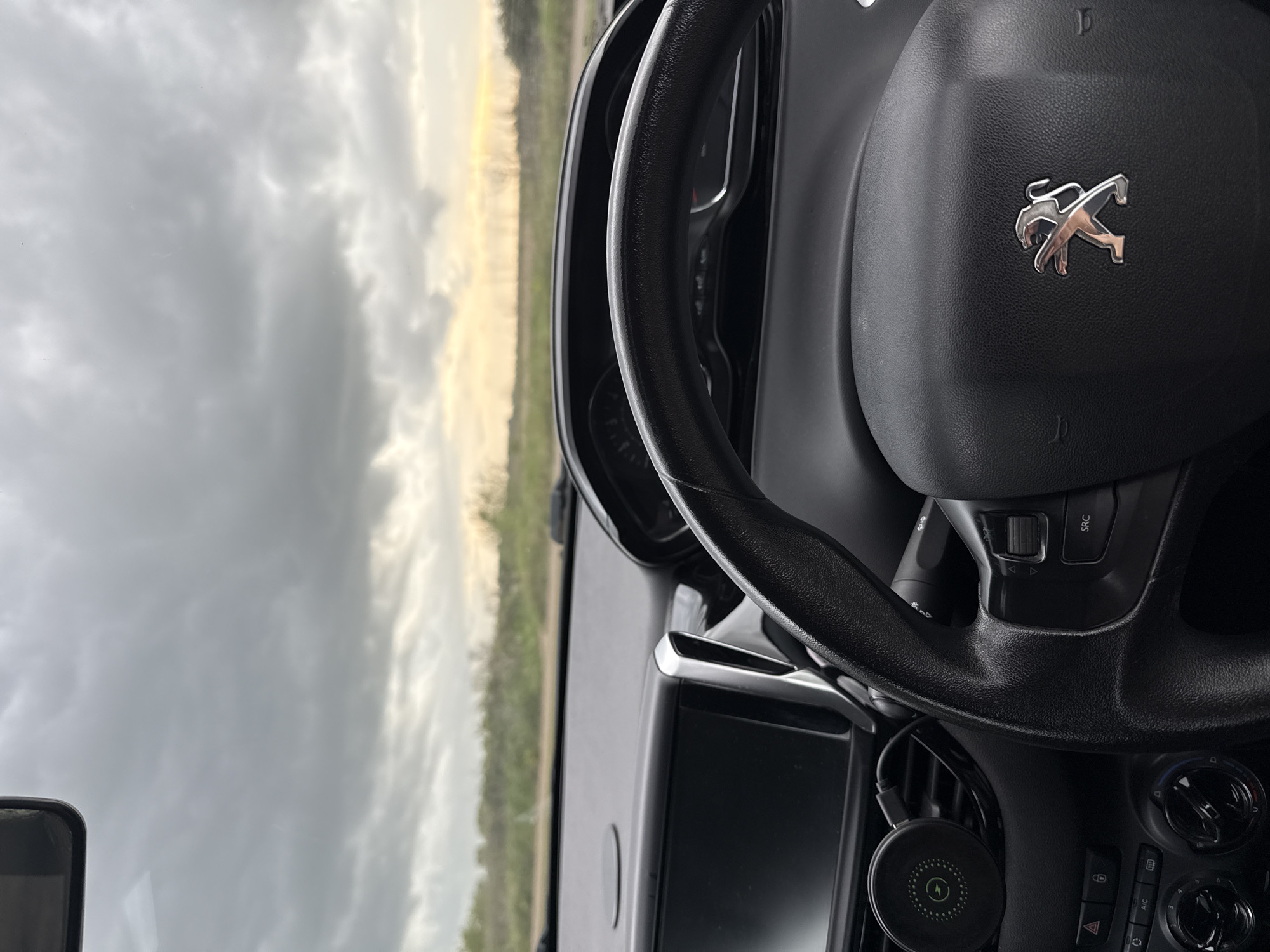 This media file captures a view from the interior of a car, looking out through the windshield towards the sky and a distant horizon. The image is presented vertically, with the car's interior details on the right and the external environment on the left.
The interior foreground is dominated by a black, textured steering wheel with a prominent silver Peugeot lion emblem at its center. Below the steering wheel, parts of the dashboard are visible, including a dark, rectangular display screen (likely an infotainment system) and a section of control buttons and dials. Visible text on these controls includes "SRC" on a steering wheel button, an "AC" button, and a triangular hazard light symbol. A black, circular device, possibly a wireless phone charger identified by a green lightning bolt symbol, is attached to an air vent on the lower dashboard.
The exterior view, seen through the windshield, shows a vast, overcast sky. Large, grey clouds dominate the scene, but a band of lighter, yellowish-white sky is visible near the distant horizon, suggesting either a break in the clouds or the soft light of dawn or dusk. A narrow strip of green land, consistent with fields or distant trees, runs along this distant horizon.
The scene depicts a moment from inside a vehicle, possibly stationary or in motion. The weather appears cloudy with diffused light. No people are visible. The context provided indicates the location is Gorleston-on-Sea, United Kingdom.