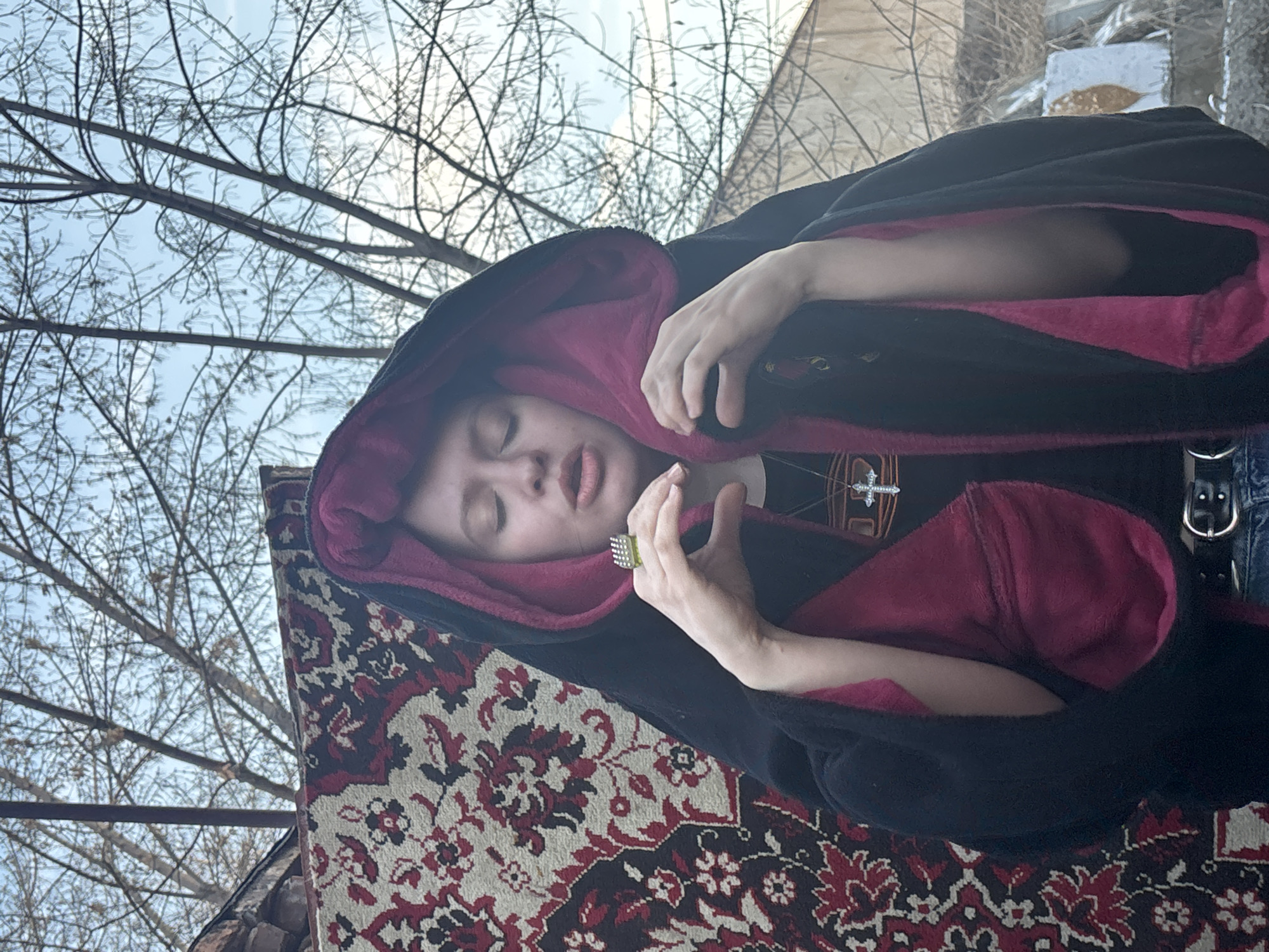 A person with closed eyes, wearing a black hooded robe with a magenta lining, is lying on a carpet with a red and cream floral pattern. The person has pale skin, a ring on their left hand, and is wearing a necklace with a cross. They are dressed in a black shirt and denim jeans, secured with a black belt with a silver buckle. The background includes bare tree branches against a blue sky and part of a building. The scene appears to be outdoors. No specific location can be identified as being in Krasnoyarsk, Russia, based solely on this image.
